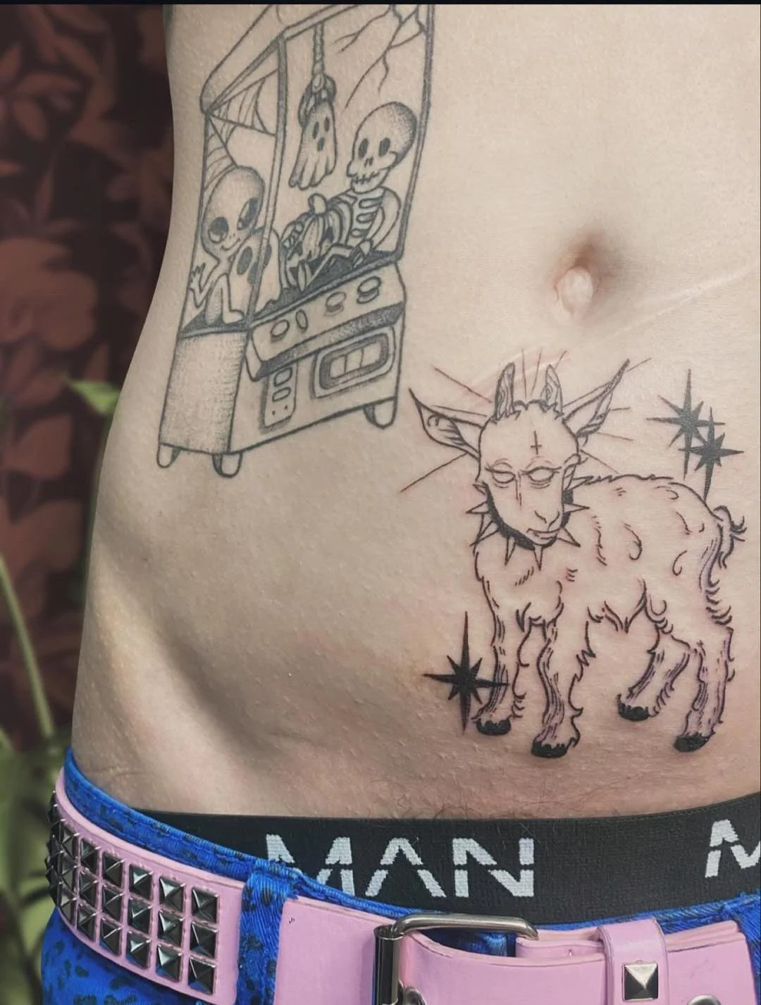 Tattoo of a goat with a cross on its forehead and star-like symbols on its body, along with a vending machine containing skeletons and a ghostly figure.