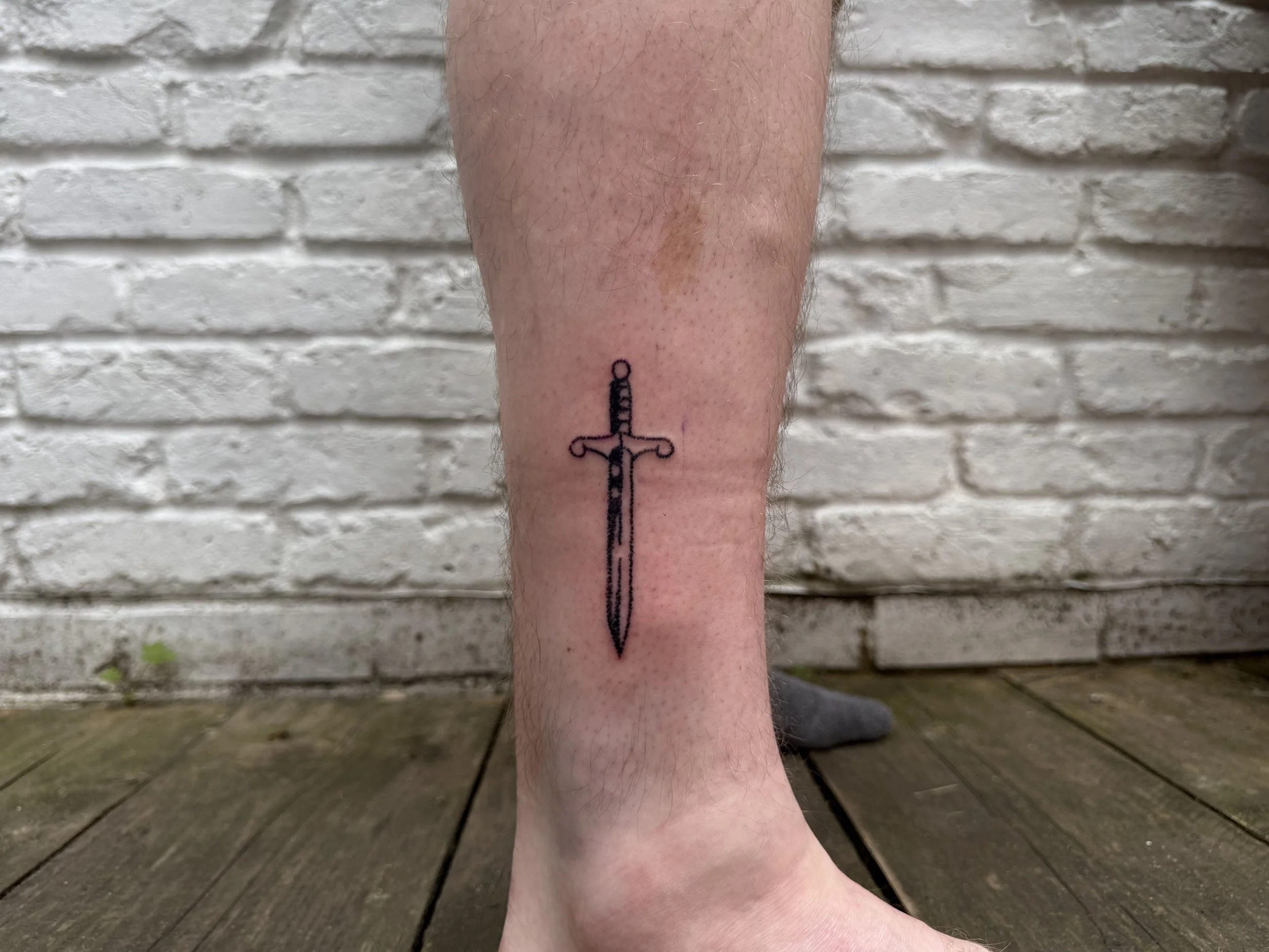 Tattoo of a black outline sword on a person's lower leg, near the ankle, with a white brick wall and wooden floor background.