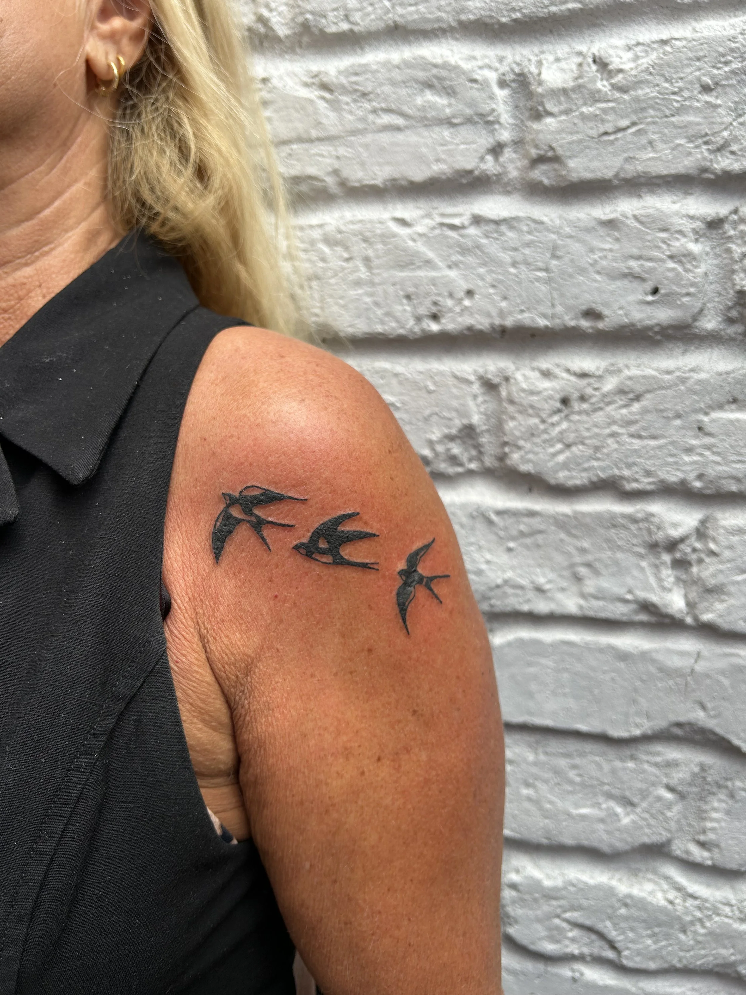 A woman with a tattoo of three black swallows flying on her shoulder against a white brick wall.