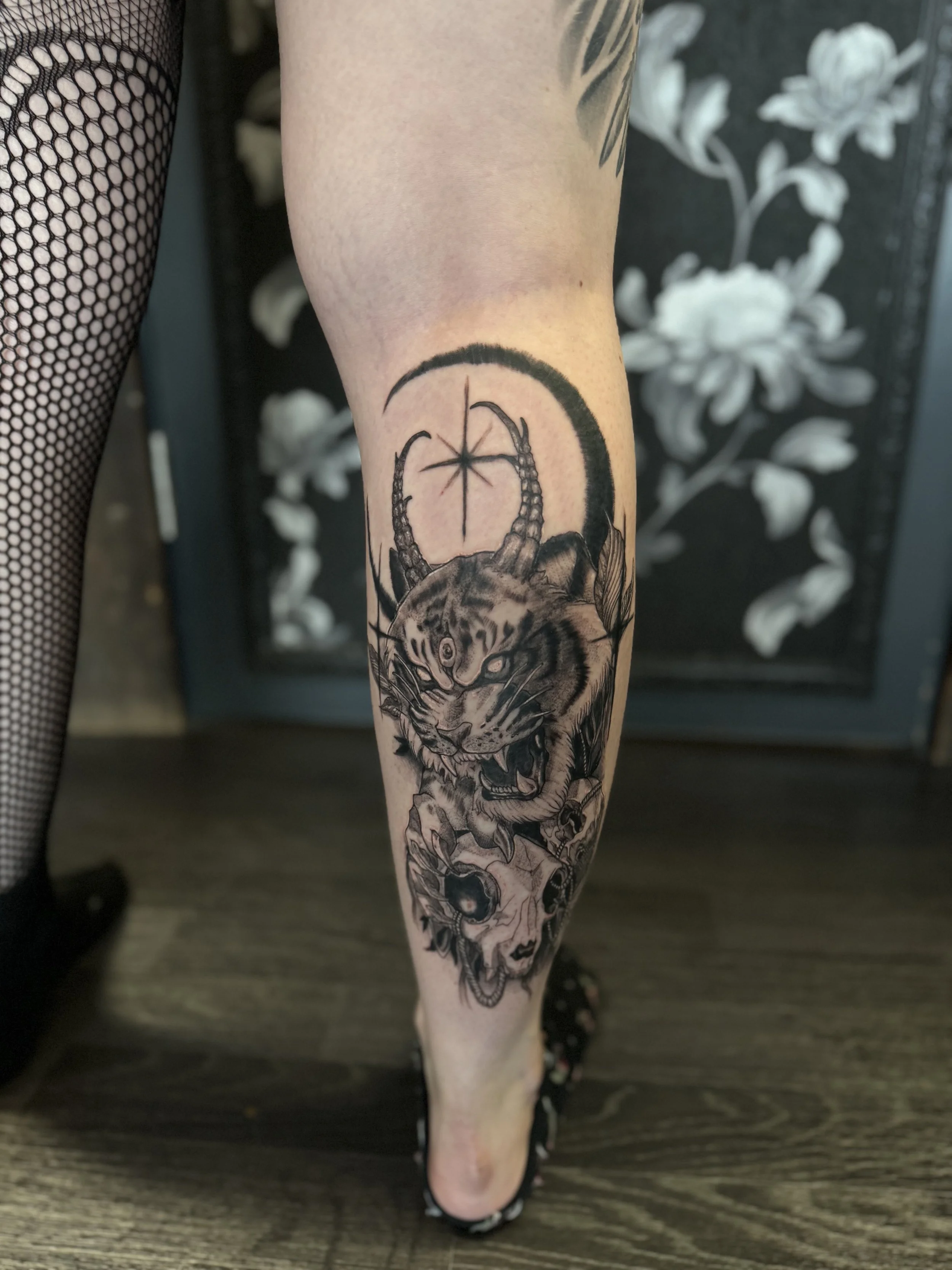 Tattoo of a fierce tiger with horns, surrounded by skulls and foliage, with a moon and star design above.