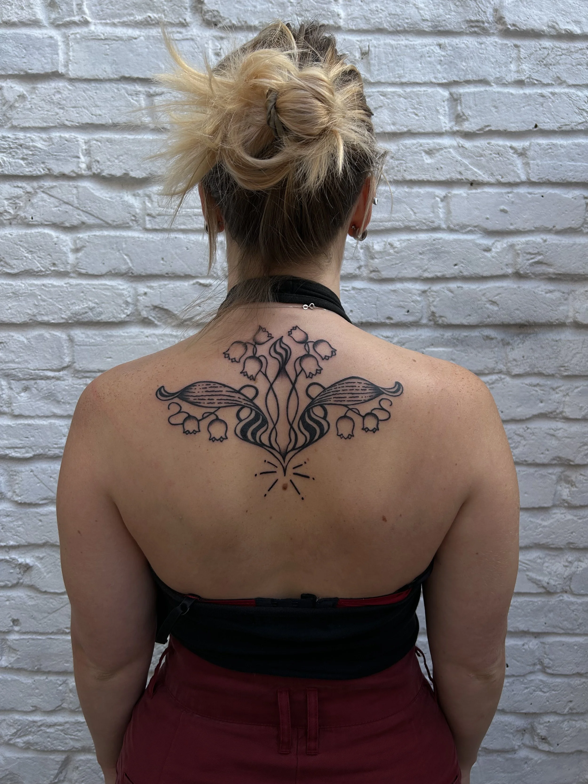A woman with blonde hair styled in a messy bun, wearing a black top and red pants, standing with her back to the camera against a white brick wall. She has a tattoo on her upper back of a symmetrical, stylized floral design with bell-shaped flowers and decorative elements.