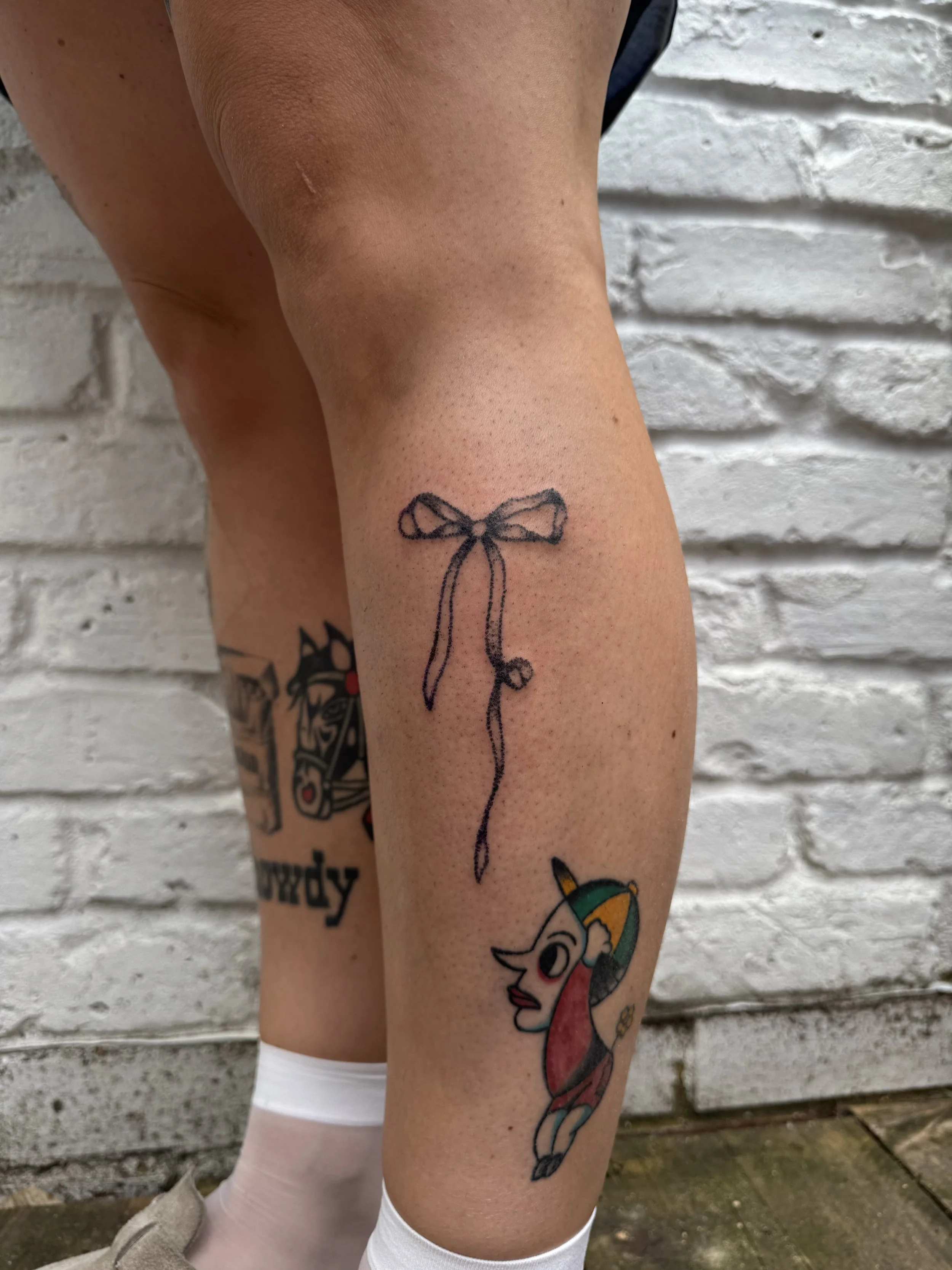 Tattoo of a knitting needle and a bow on a person's calf, with another colorful cartoon unicorn tattoo below.