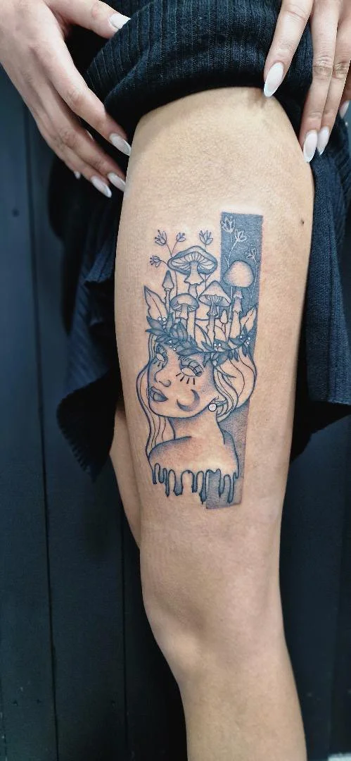 Tattoo of a woman's face with long hair, surrounded by mushrooms and plants, with a large moon in the background.