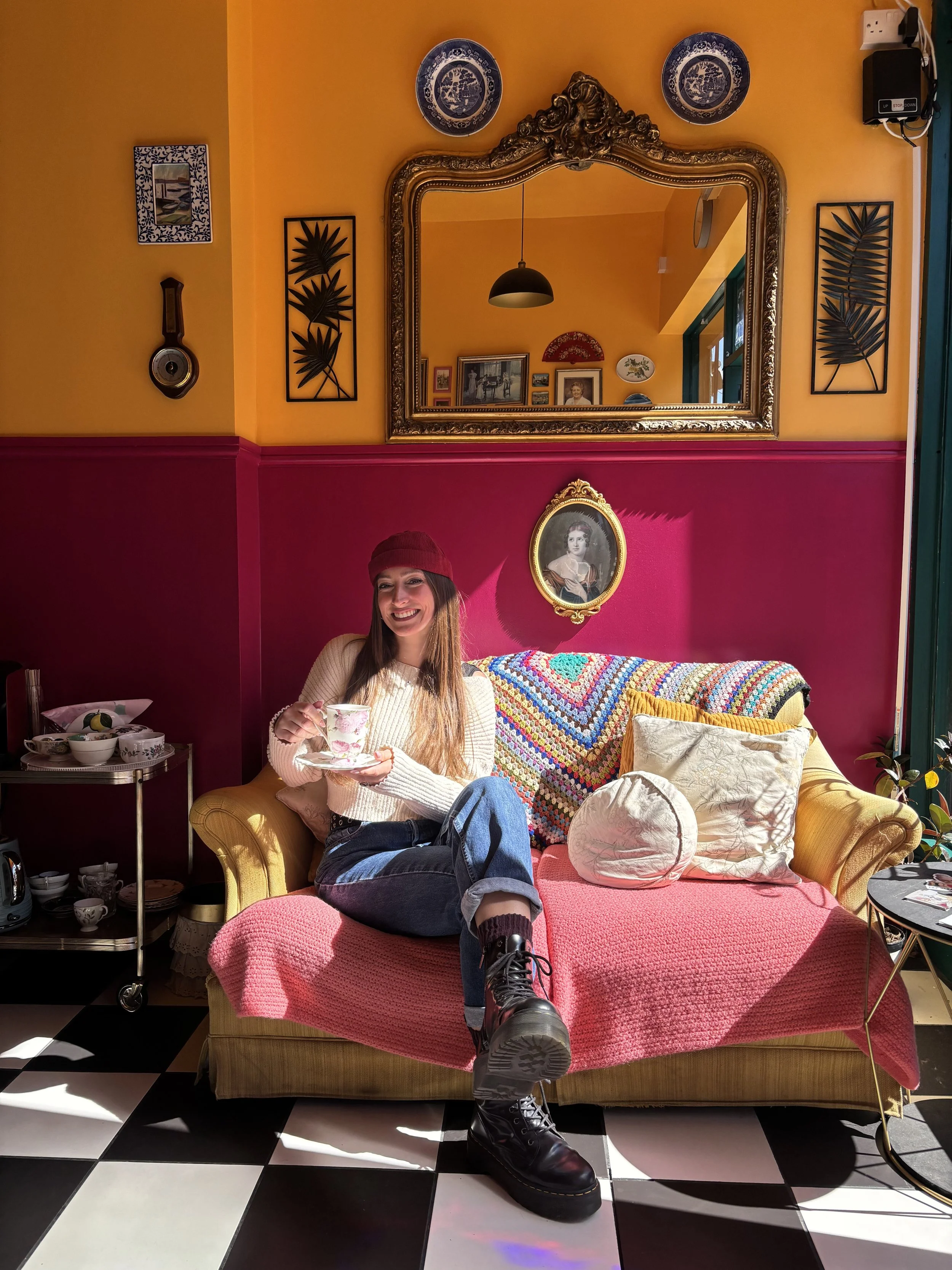 A woman with long brown hair wearing a red beanie, white sweater, and black boots, sitting on a colorful pink and yellow sofa in a room with yellow walls and a black and white checkered floor. She is smiling and holding a teacup and saucer. Behind her, the wall has various framed pictures, a decorative portrait, and two bookshelves with cups and glasses. A large ornate gold-framed mirror hangs above the sofa, reflecting the room's interior.