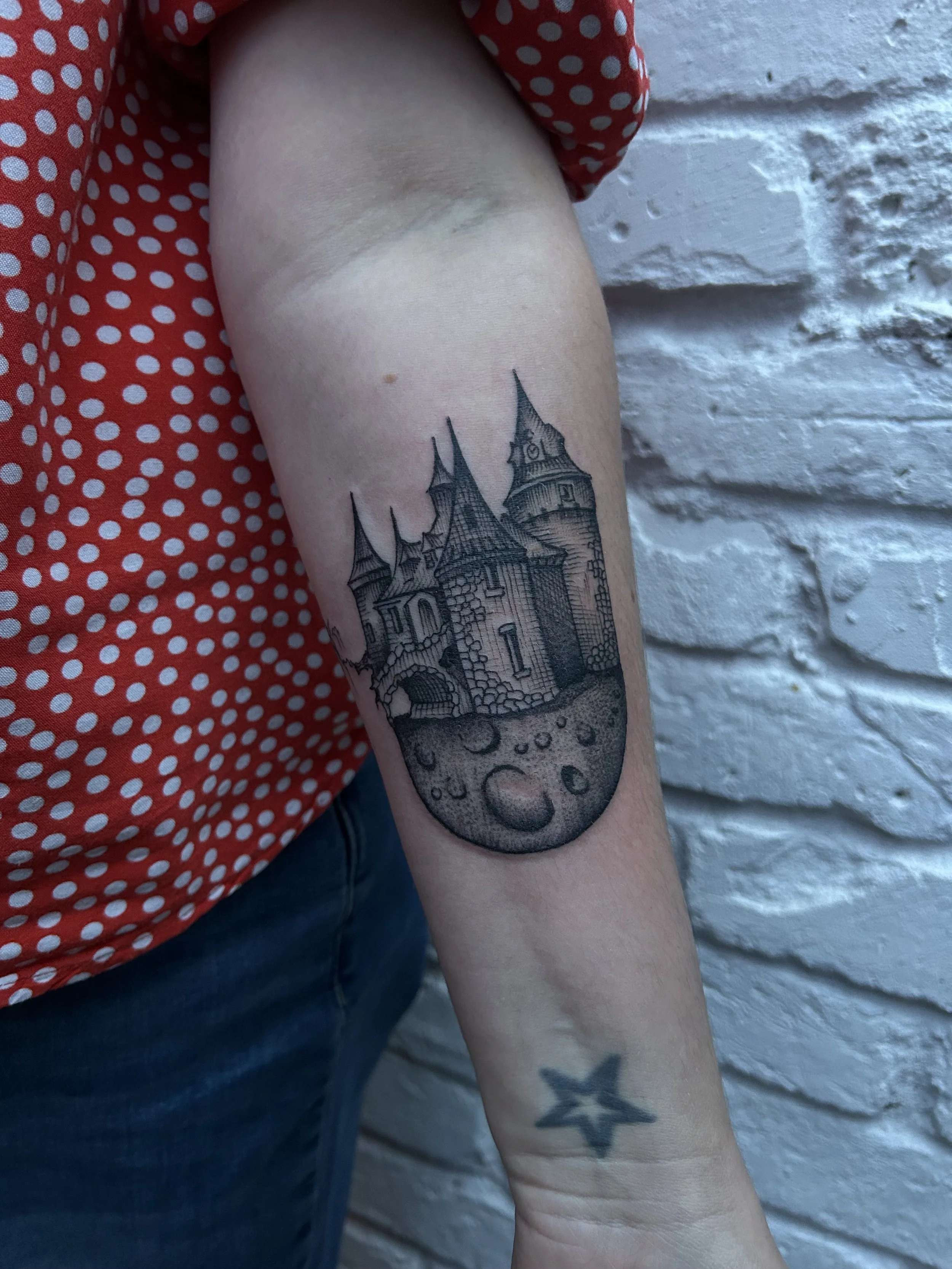 Tattoo of a castle with towers and turrets, with moon and crater details underneath, on a person's upper arm.