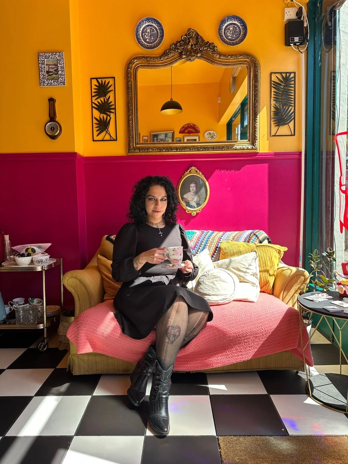 Woman sitting on a colorful vintage sofa in a cafe or tea room, holding a teacup and saucer. The room features a vibrant pink and yellow wall, decorative plates, framed pictures, and a large ornate mirror. The floor has a black and white checkered pattern, and sunlight streams in from a nearby window.