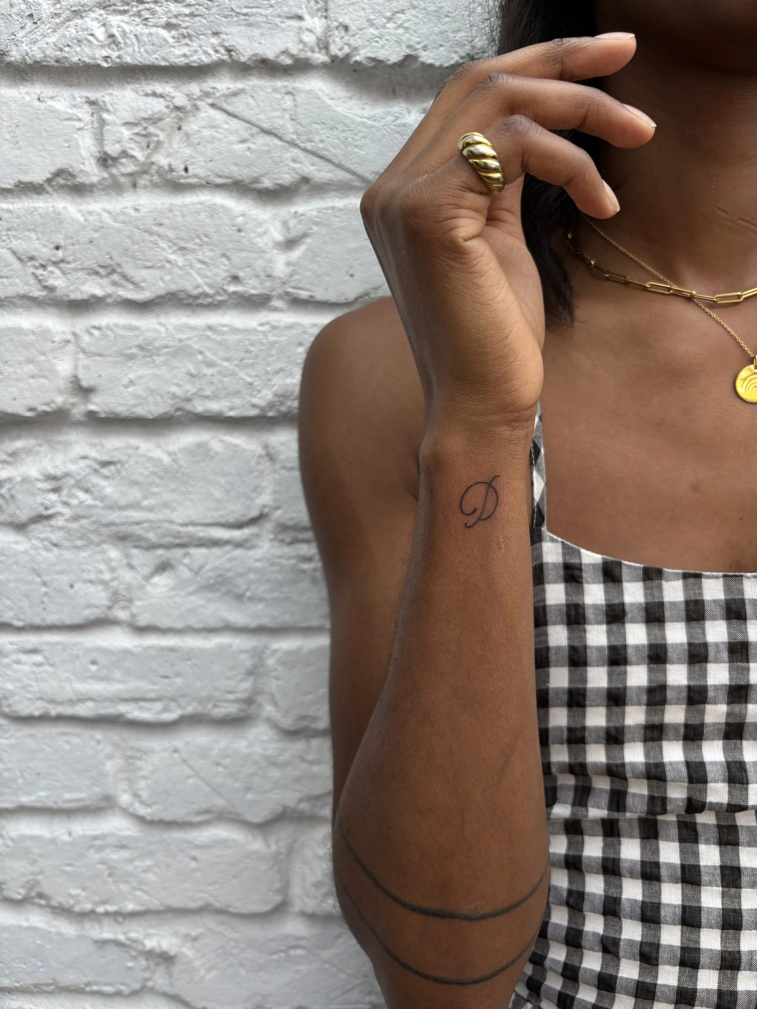 Close-up of a woman’s arm with a small black script letter 'D' tattoo on her wrist, her hand near her face, wearing a gold ring and layered gold necklaces, against a white brick wall. She is dressed in a black and white checkered sleeveless top.