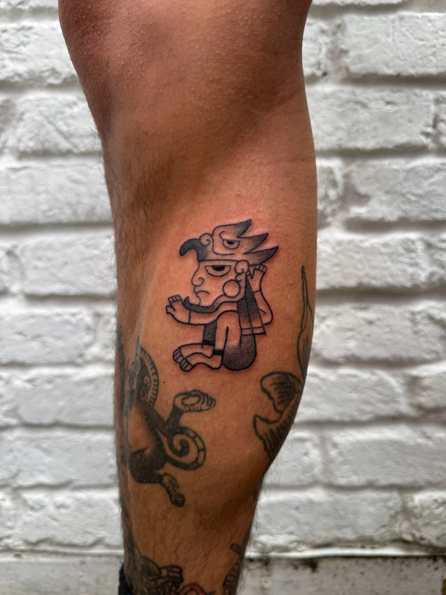 Tattoo of a cartoonish creature with a human face, animals, and abstract features on a person's arm, in front of a white brick wall.