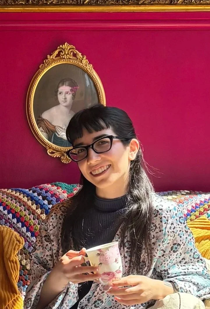 A young woman with dark hair, glasses, and a patterned cardigan smiling and holding a floral mug, sitting on a colorful crocheted blanket in front of a red wall with a gold-framed portrait of a woman.
