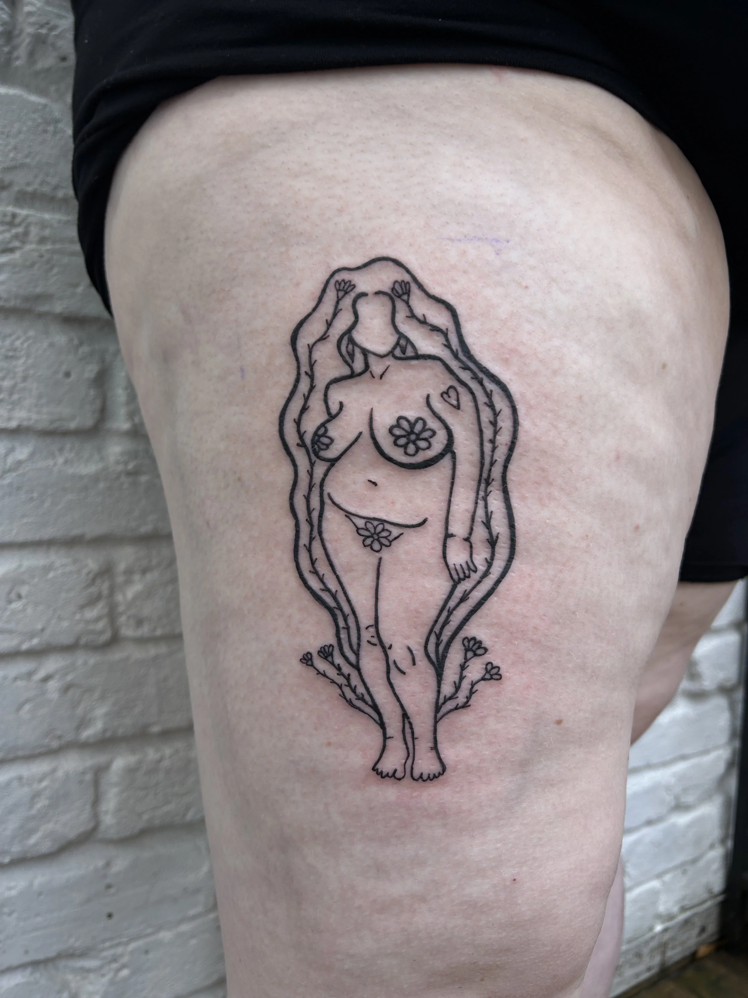 Tattoo of a woman with long hair, flowers covering the chest and private areas, and surrounded by a vine, on a person's thigh.