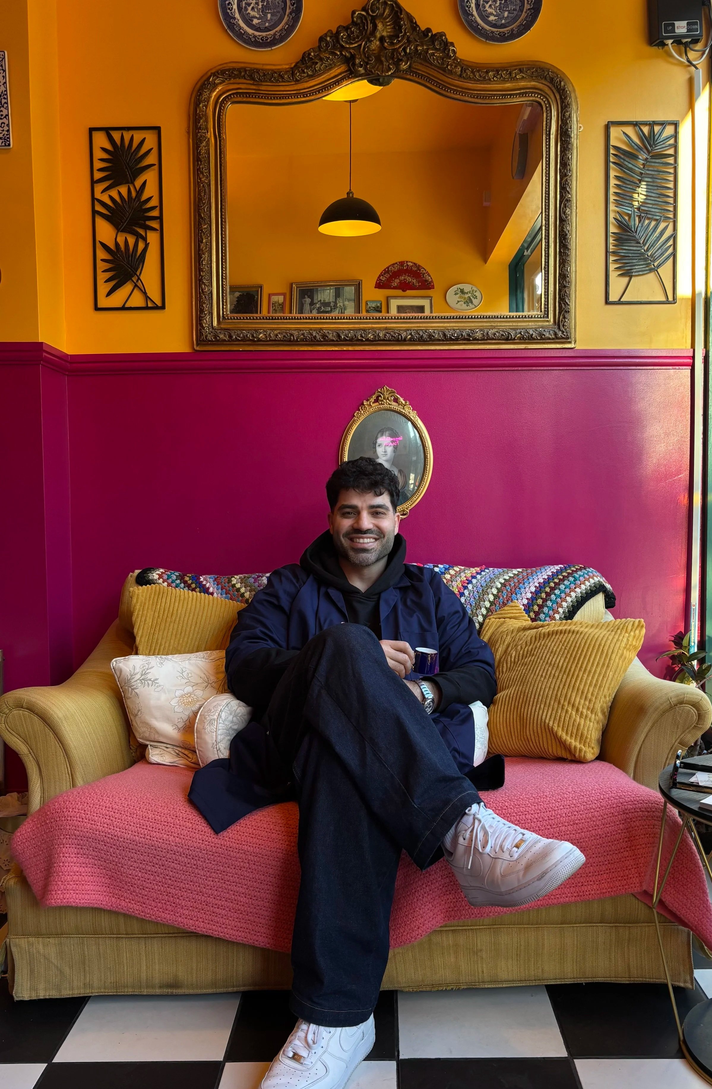 A man sitting on a vintage yellow sofa with a pink cover, smiling and holding a small cup. The room has bright yellow and pink walls, decorative frames, and a large ornate mirror above the sofa, reflecting a hanging light and art on the opposite wall.
