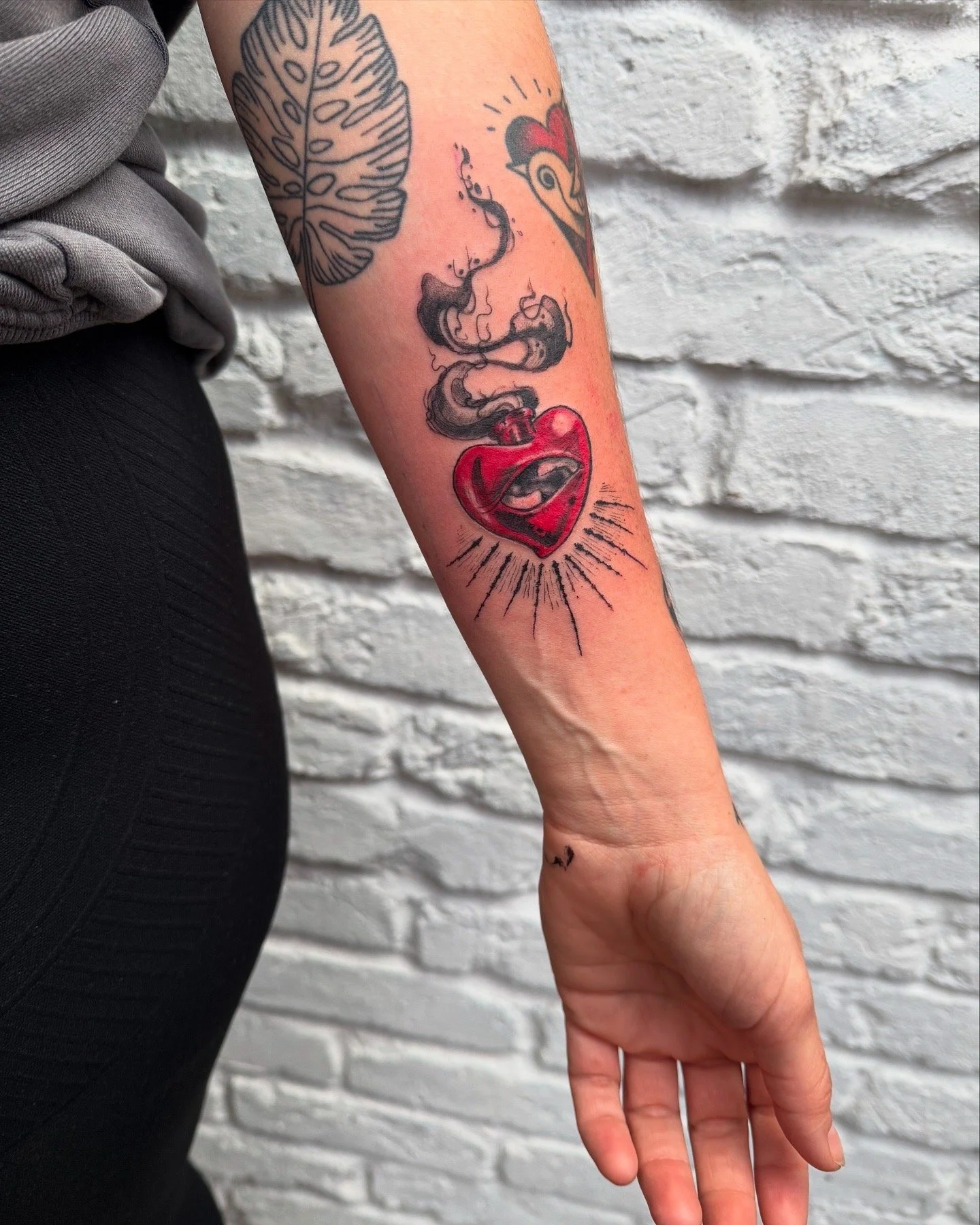 Tattoo of a red heart with a crack, from which smoke rises, on the person's forearm.