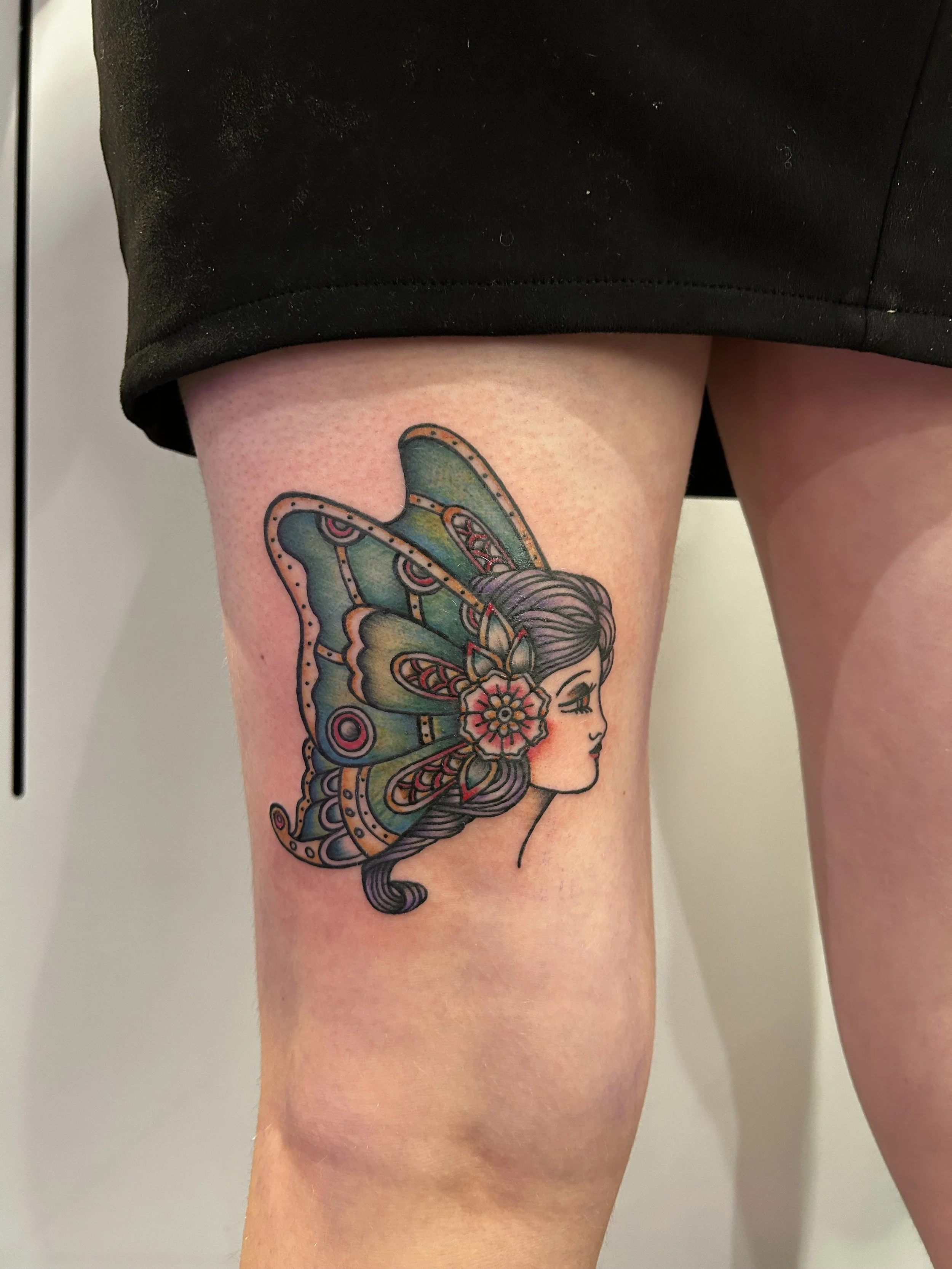 Colorful tattoo of a woman's profile with butterfly wings on her head, adorned with a flower, on a person's thigh.
