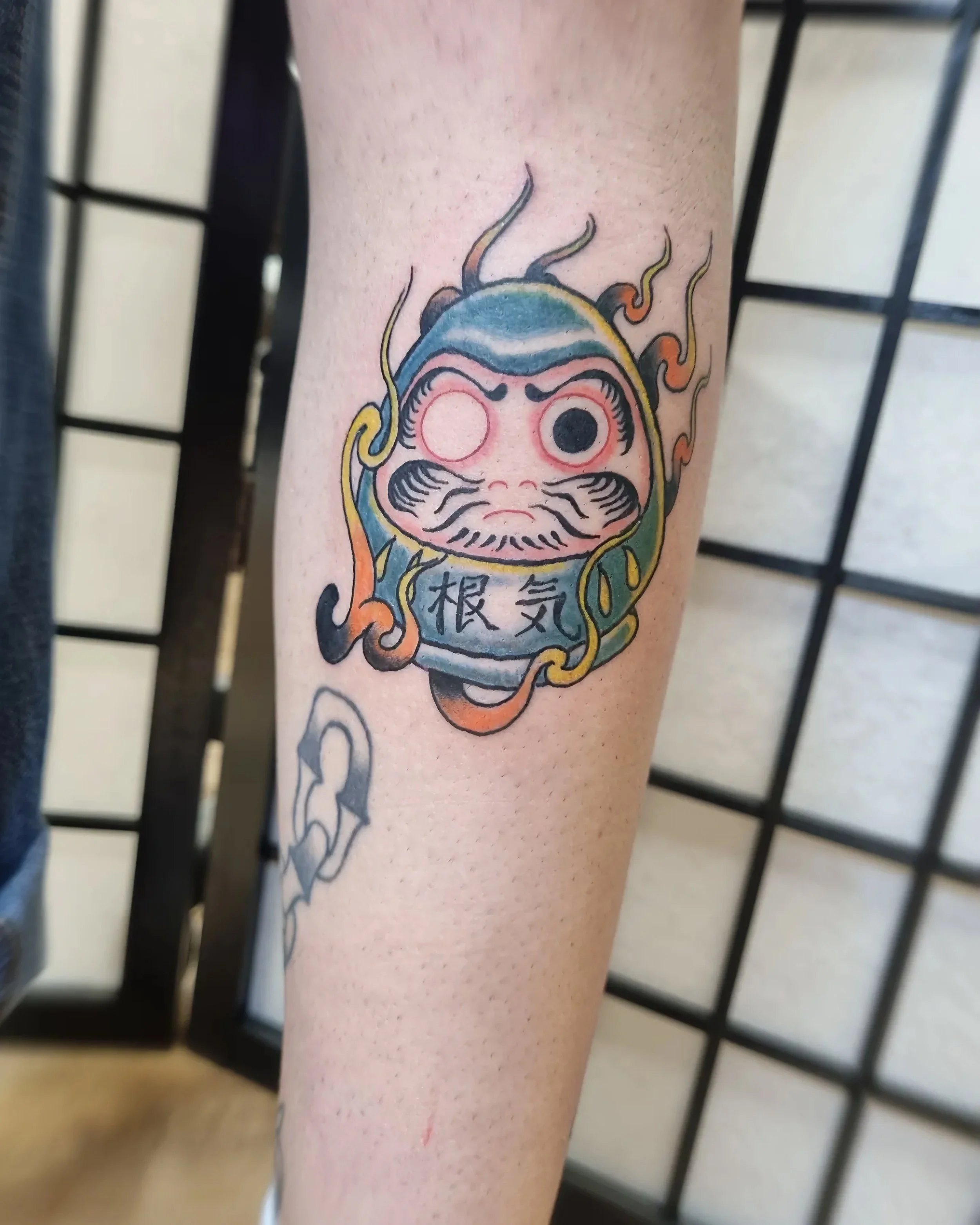 Tattoo of a Daruma doll with one eye filled in, surrounded by flames and Japanese characters.