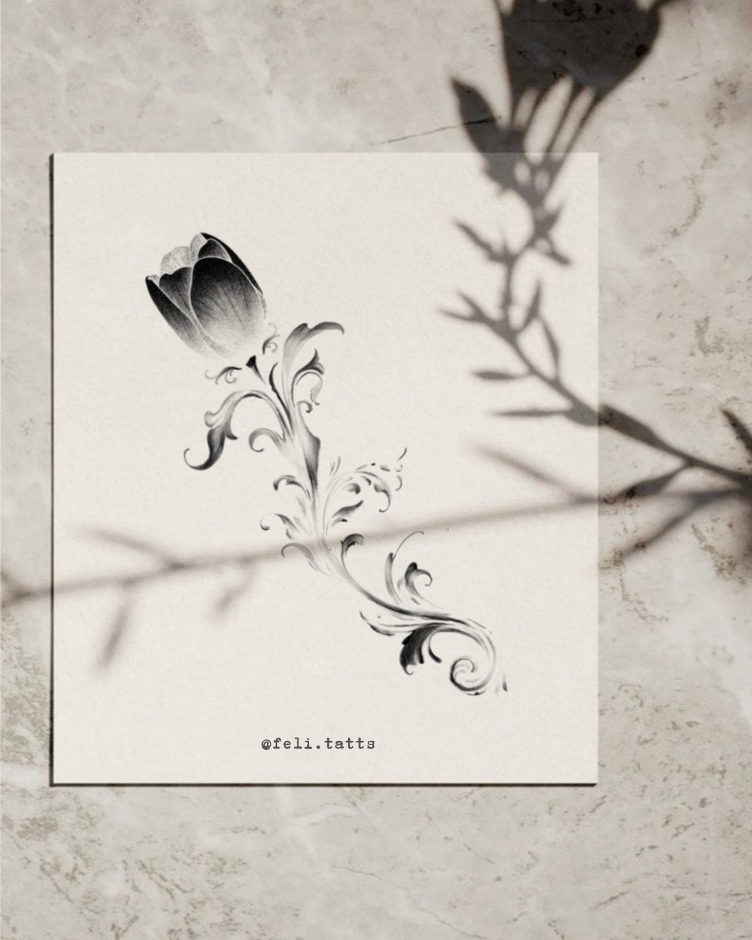 A black and white floral tattoo design of a single tulip with swirling decorative leaves on white paper. The paper has the text '@feli.tatts' at the bottom. Shadows of a plant cast across the paper and surface.
