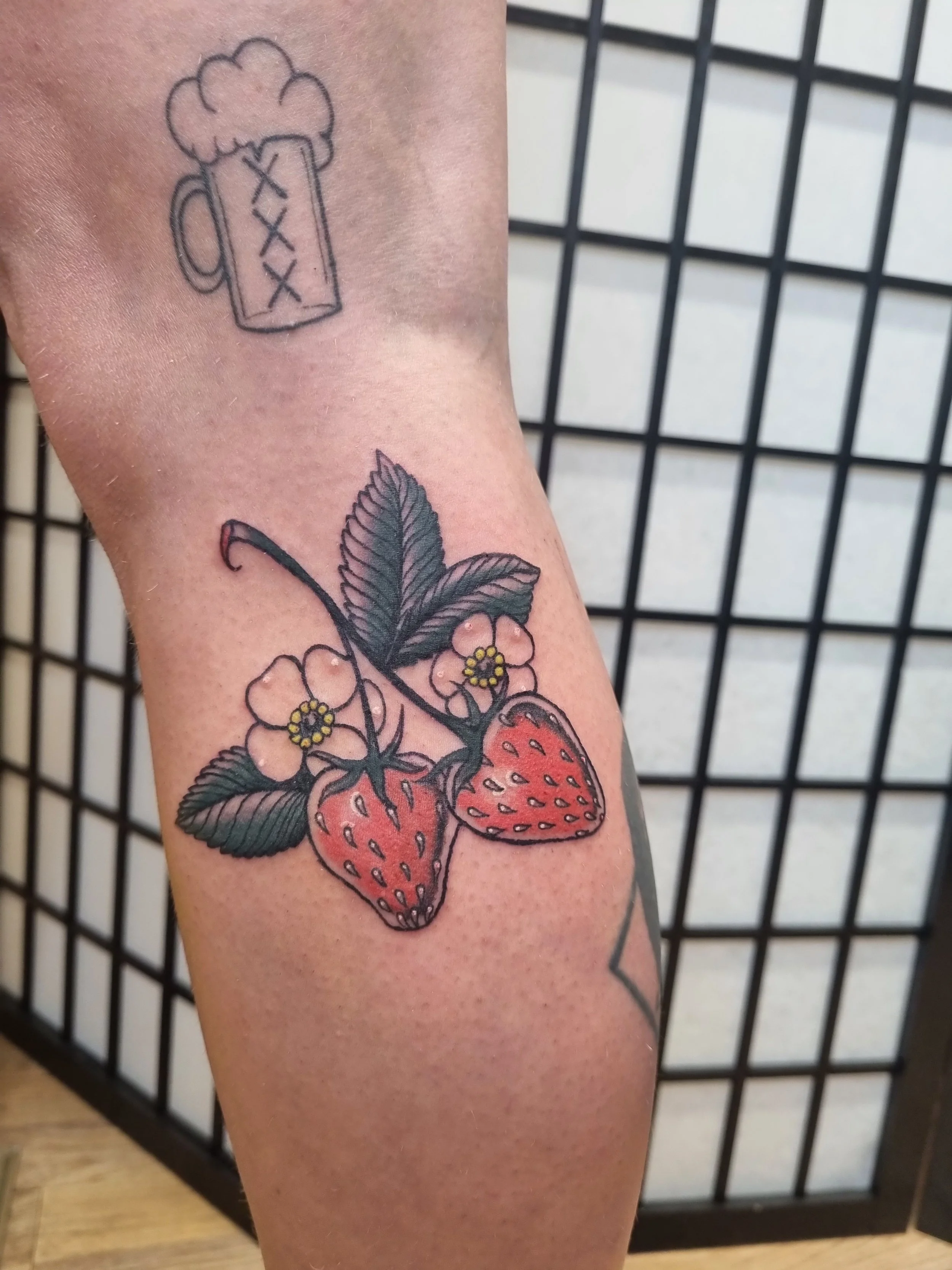 Tattoo of a strawberry plant with two strawberries, pink flowers, and green leaves on a person's forearm, with a smaller, simpler beer mug tattoo above it.