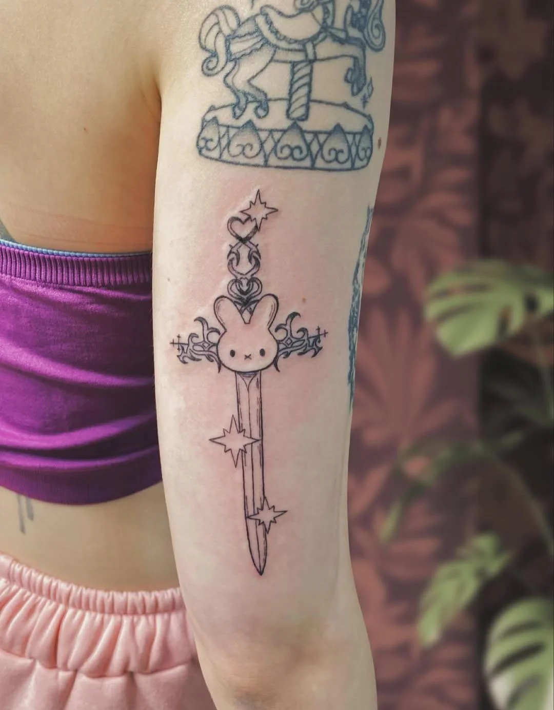 Tattoo of a cartoon bunny with a sword, stars, and decorative flourishes on a person's forearm.