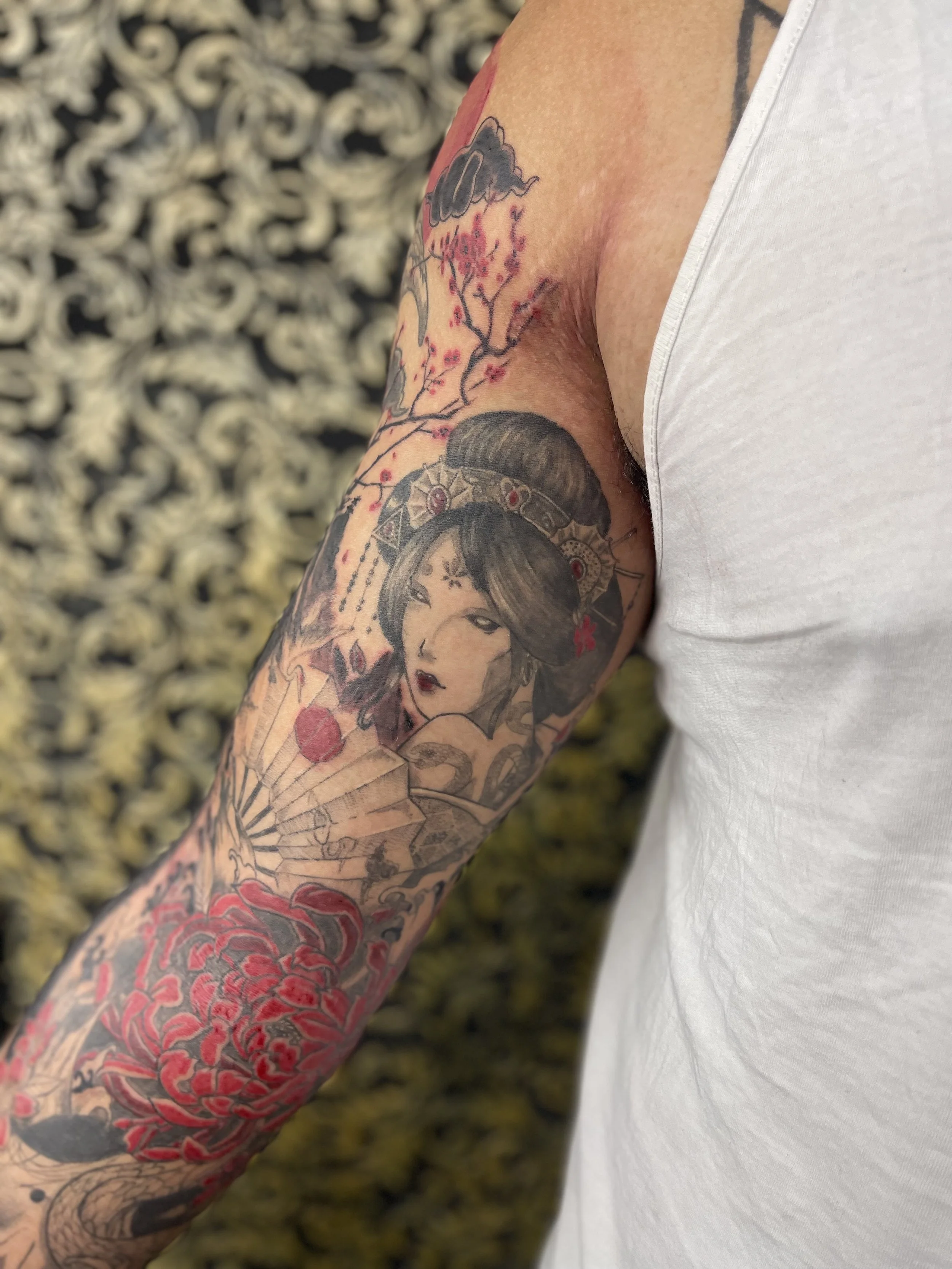 Tattoo sleeve featuring a woman with traditional Asian hairstyle, cherry blossoms, a fan, a snake, and vibrant red flowers.