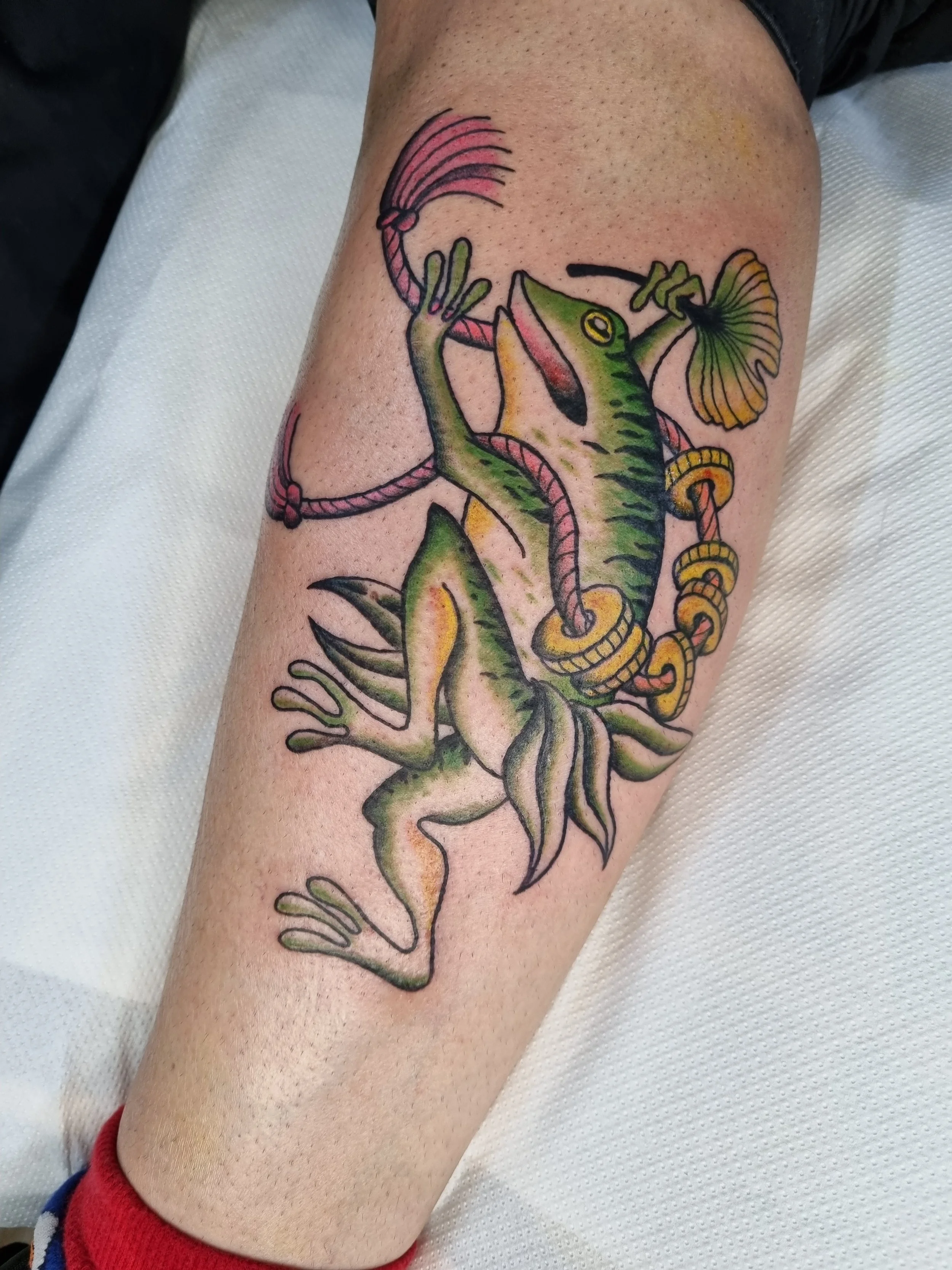Colorful tattoo of a chameleon holding a paintbrush, wearing jewelry, with a skeleton tail, on a person's leg.
