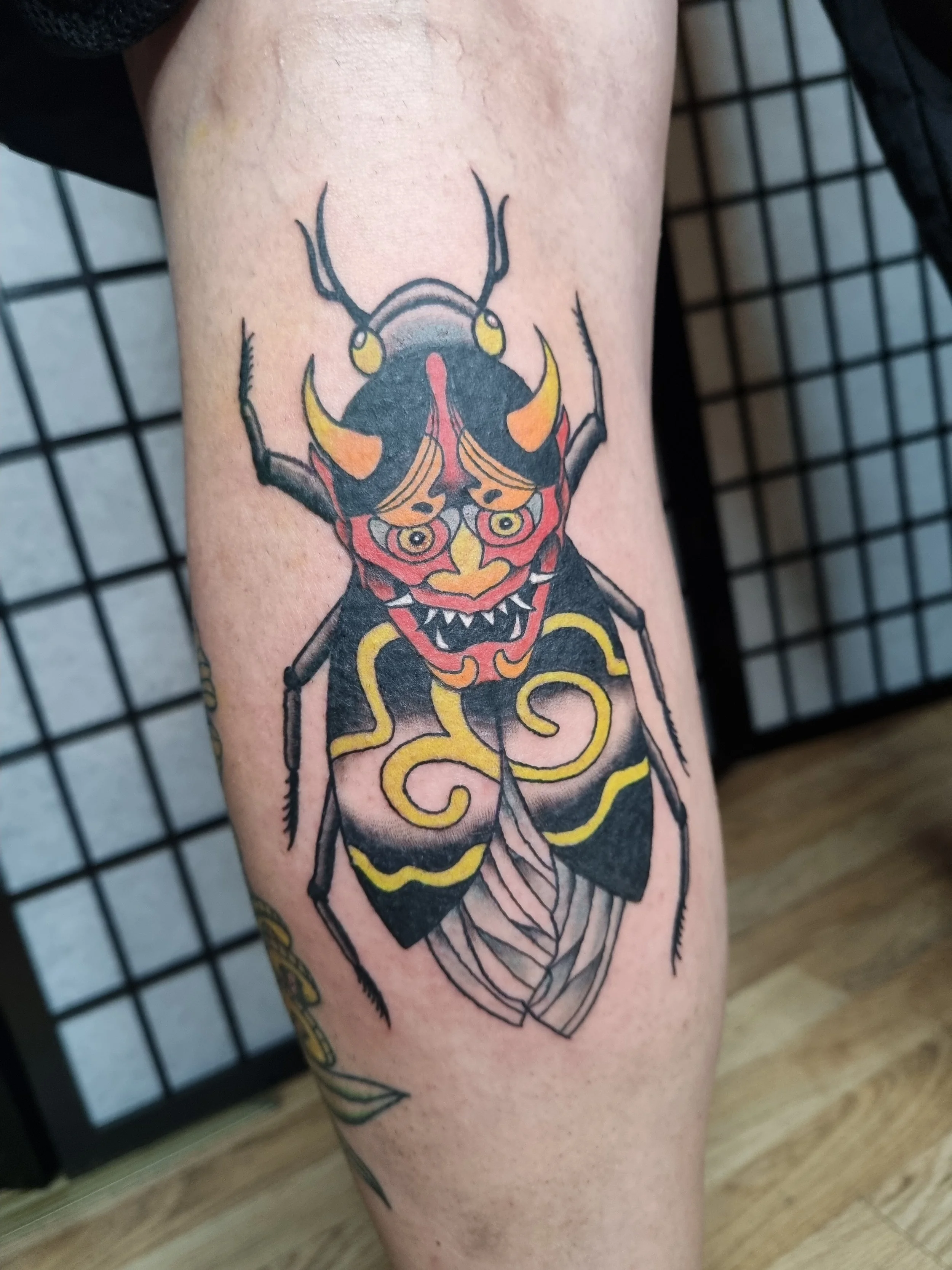 Tattoo of a stylized insect with a fierce face, colorful yellow, red, and black markings, and swirling details on its body