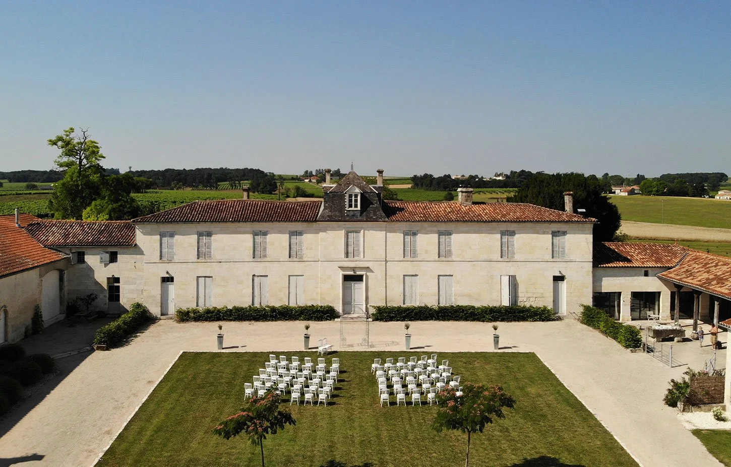 A large historic building with a red tile roof, white walls, and multiple windows, set in a rural landscape with green fields. In front, an outdoor wedding ceremony setup with white chairs arranged on a grassy area, surrounded by a clear, sunny sky.
