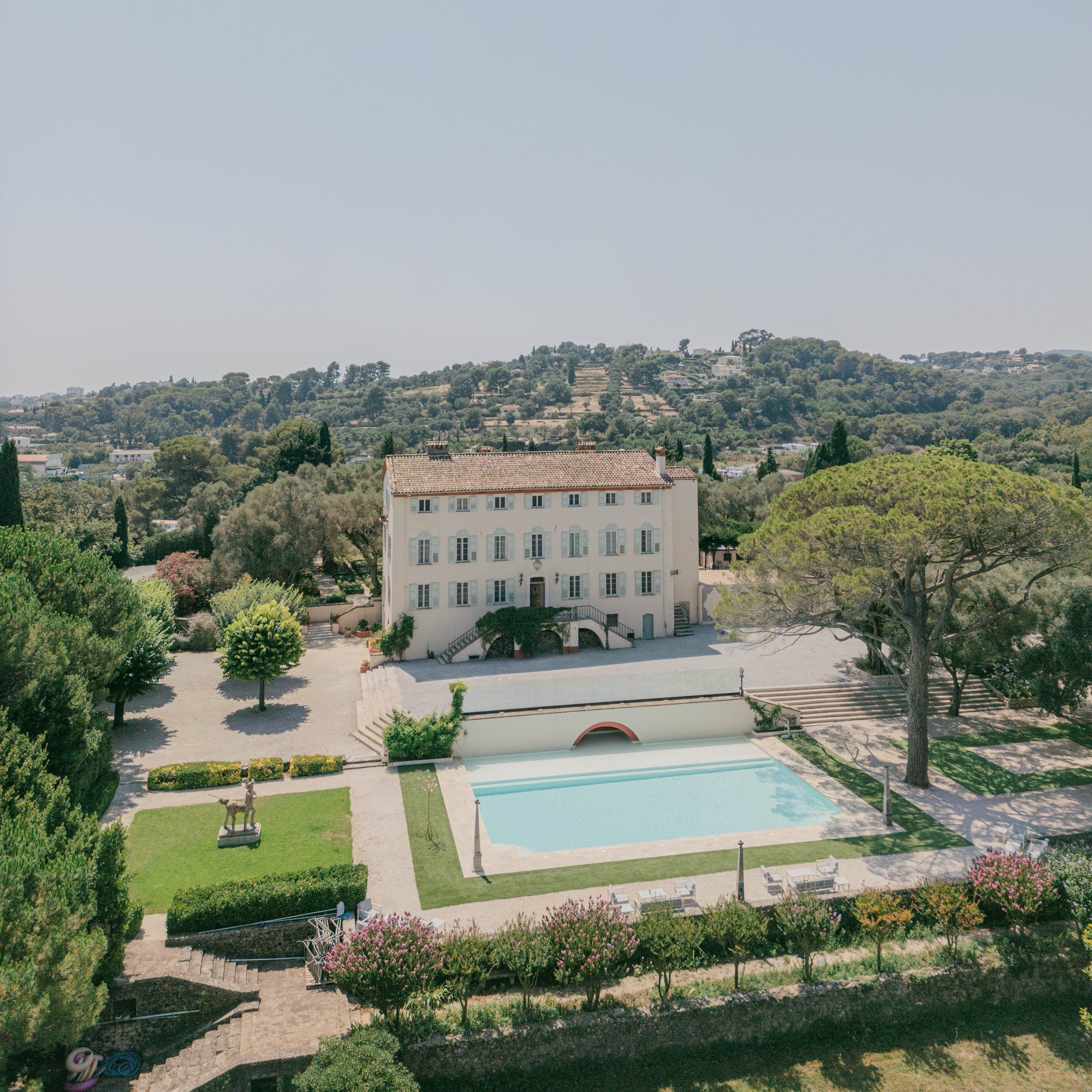 A large white historic villa with a tiled roof, set amidst lush greenery and trees, with a swimming pool and outdoor seating area in the foreground, and rolling hills in the background on a bright day.