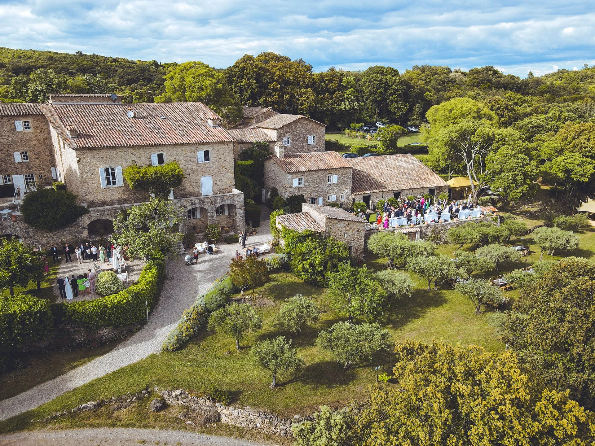 Aerial view of a rustic stone building estate during a gathering event, surrounded by green trees and landscaped gardens.