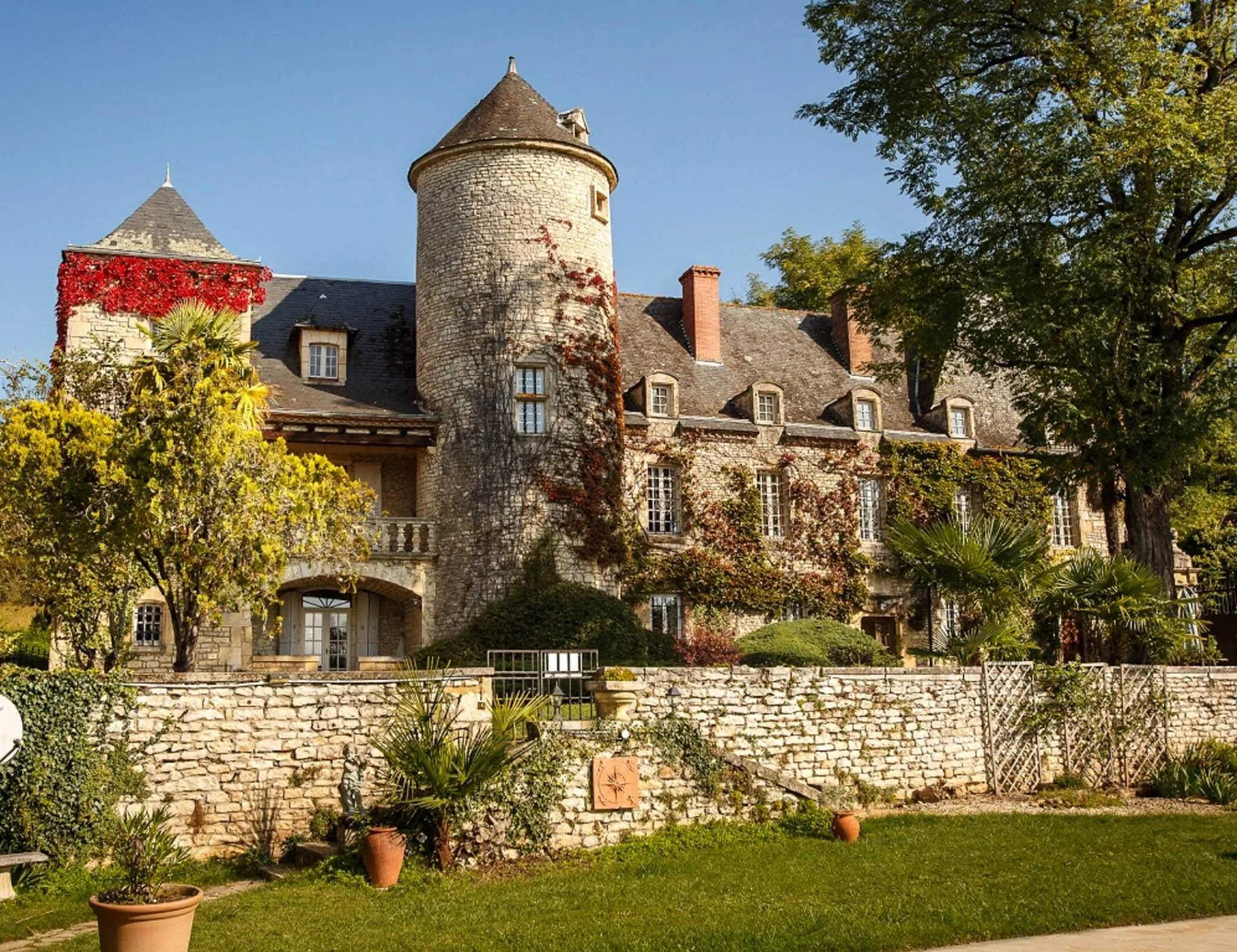 A stone castle with a turret, surrounded by greenery, trees, and a stone wall in the foreground.