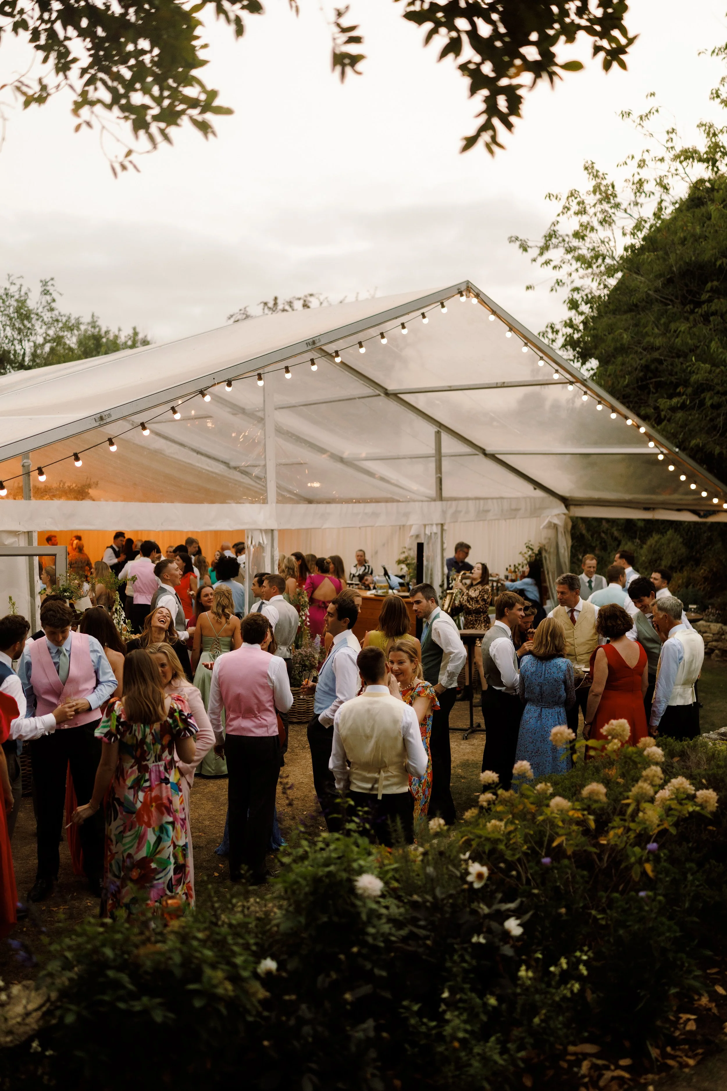 Greenhouse marquee wedding reception with festoon lighting on the trim of the structure. Guests all mingle outside for cocktail hour whilst a DJ sax plays