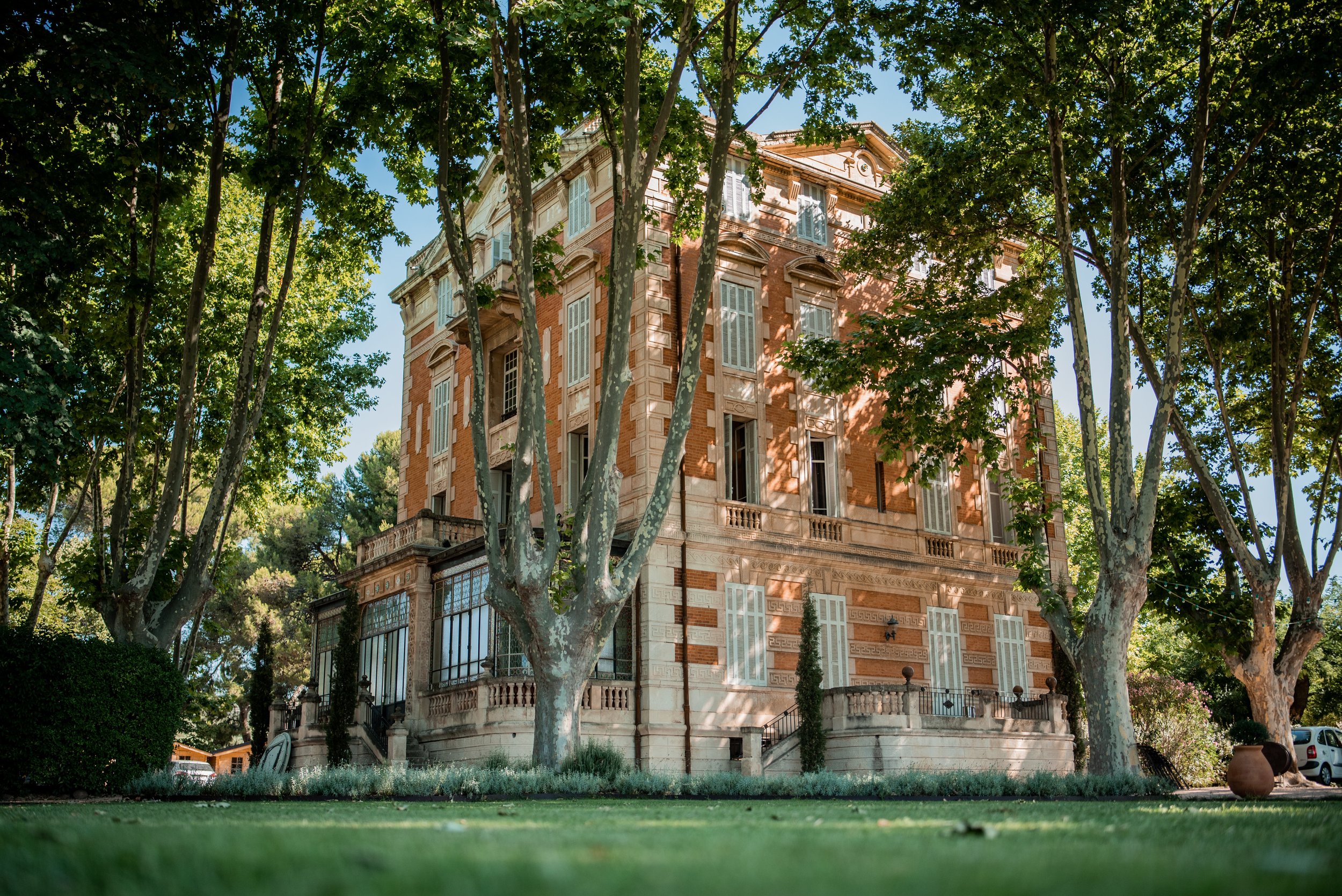 A historic brick mansion with large windows, surrounded by trees with green leaves, set on a well-maintained lawn.