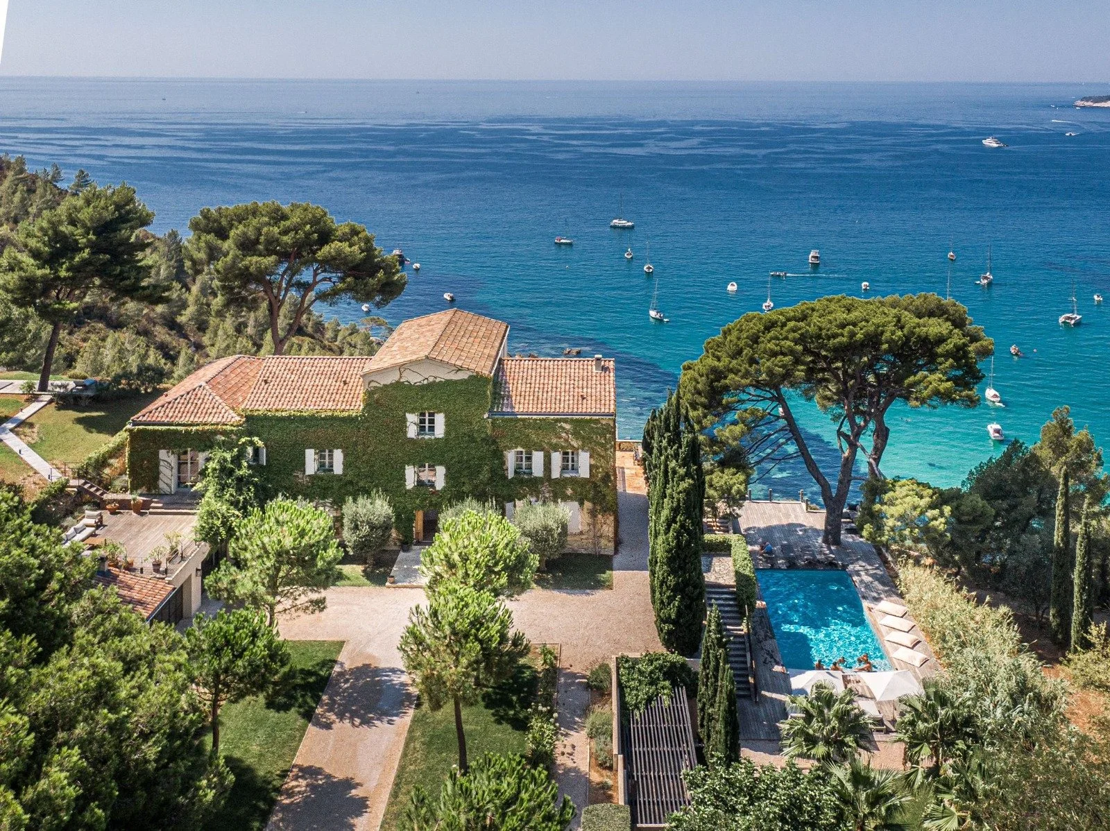 Aerial view of a luxurious house with a swimming pool, surrounded by lush trees, overlooking the ocean with boats anchored in the water.