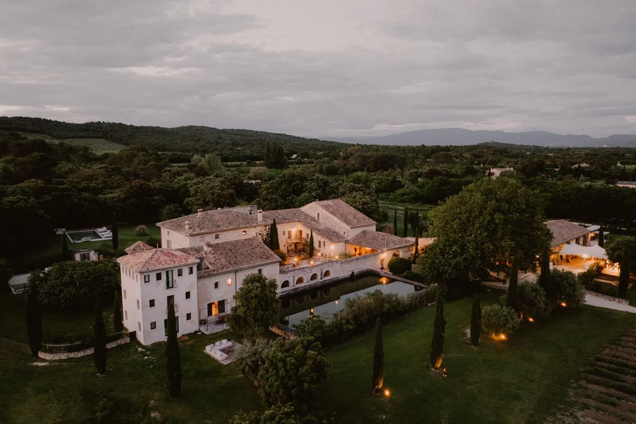 Aerial view of a large estate with multiple buildings, a pond, and surrounding lush greenery at dusk.