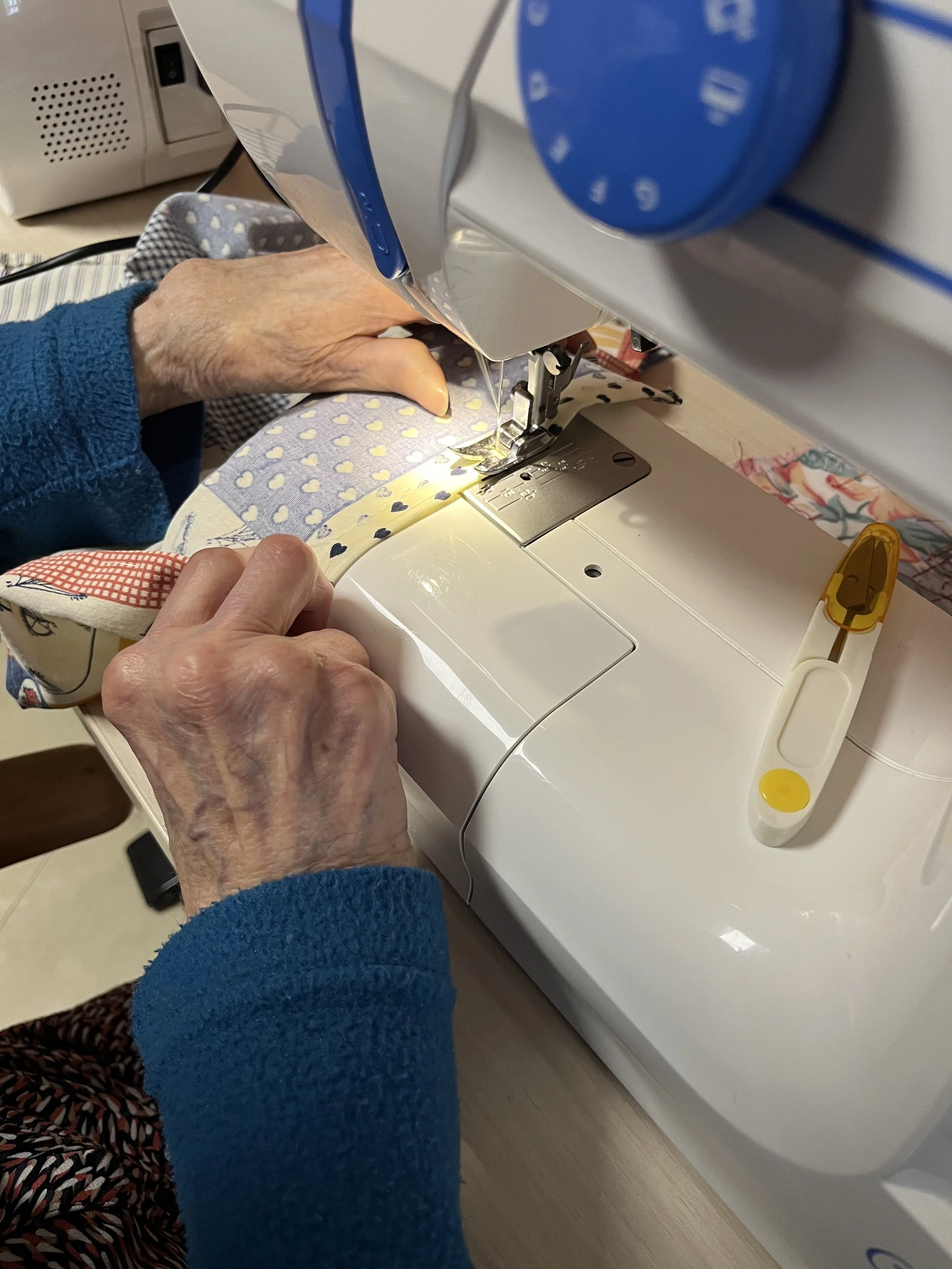Une personne âgée utilisant une machine à coudre pour faire de la couture. La machine est blanche avec quelques éléments jaunes et un petit appareil à côté.