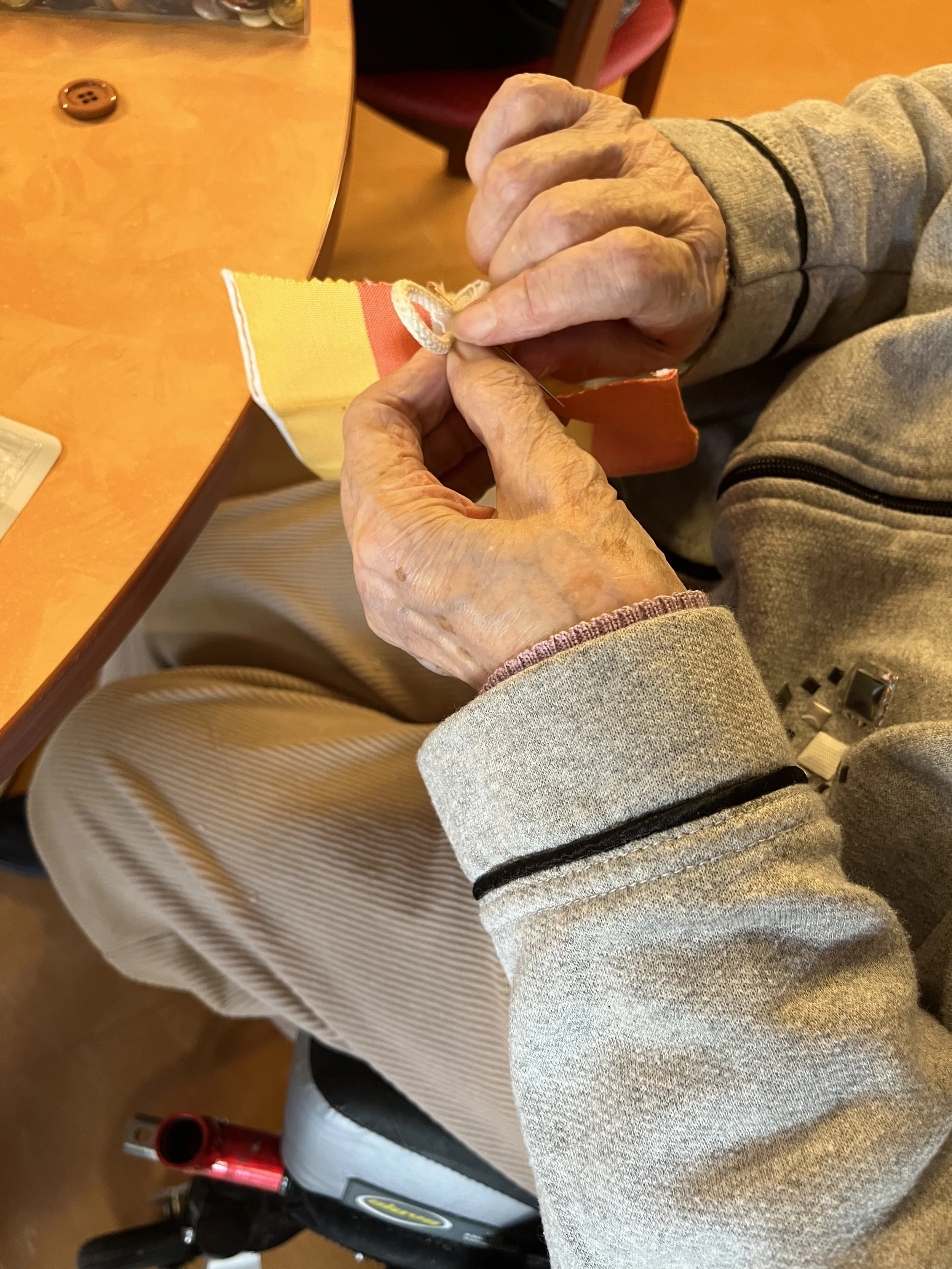Mains d'une personne cousant un tissu multicolore, assise à une table en bois.