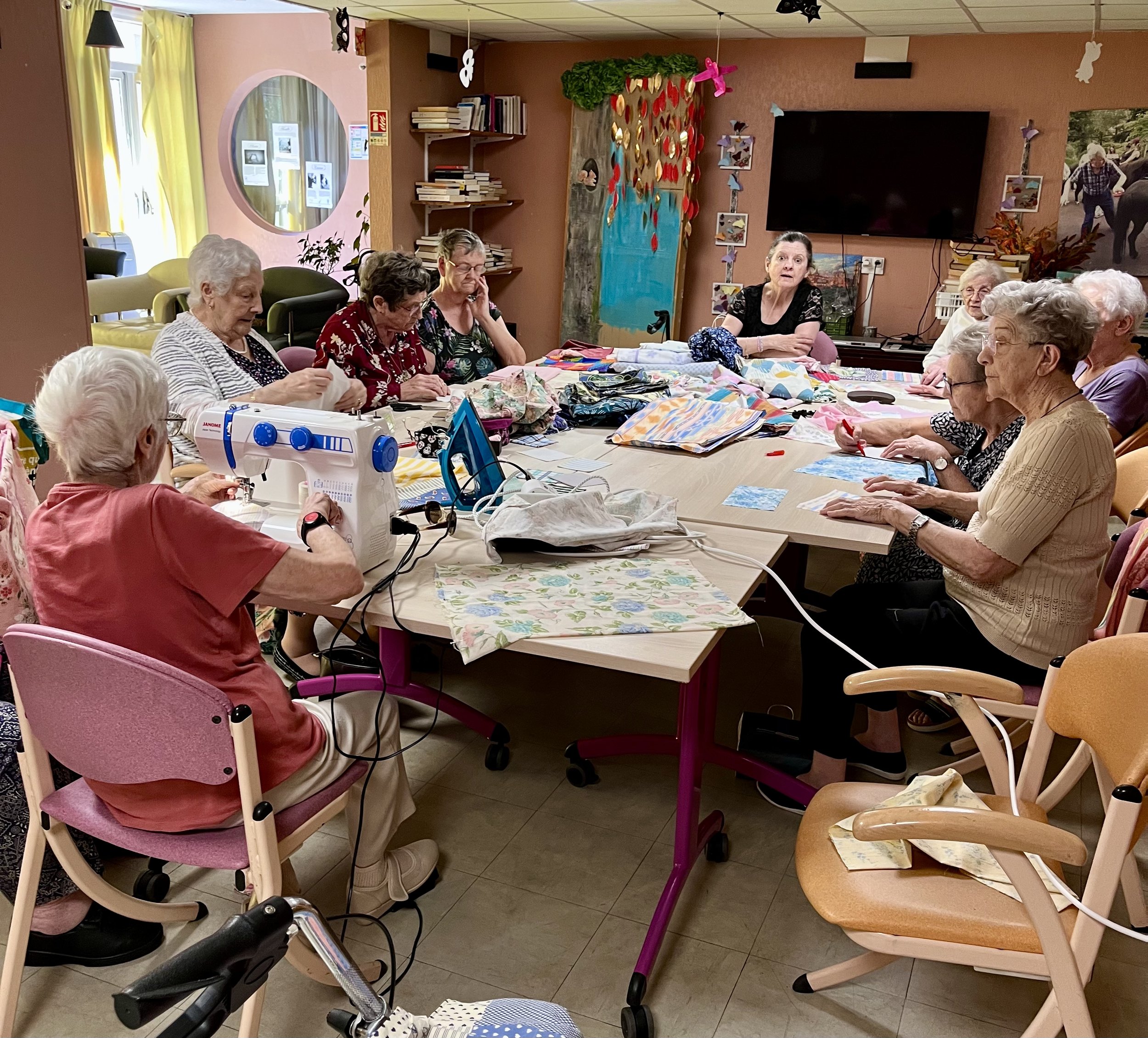 Groupe de personnes âgées réunies autour d'une table pour une activité de couture ou de tricot dans une salle décorée avec des œuvres artistiques et des livres.