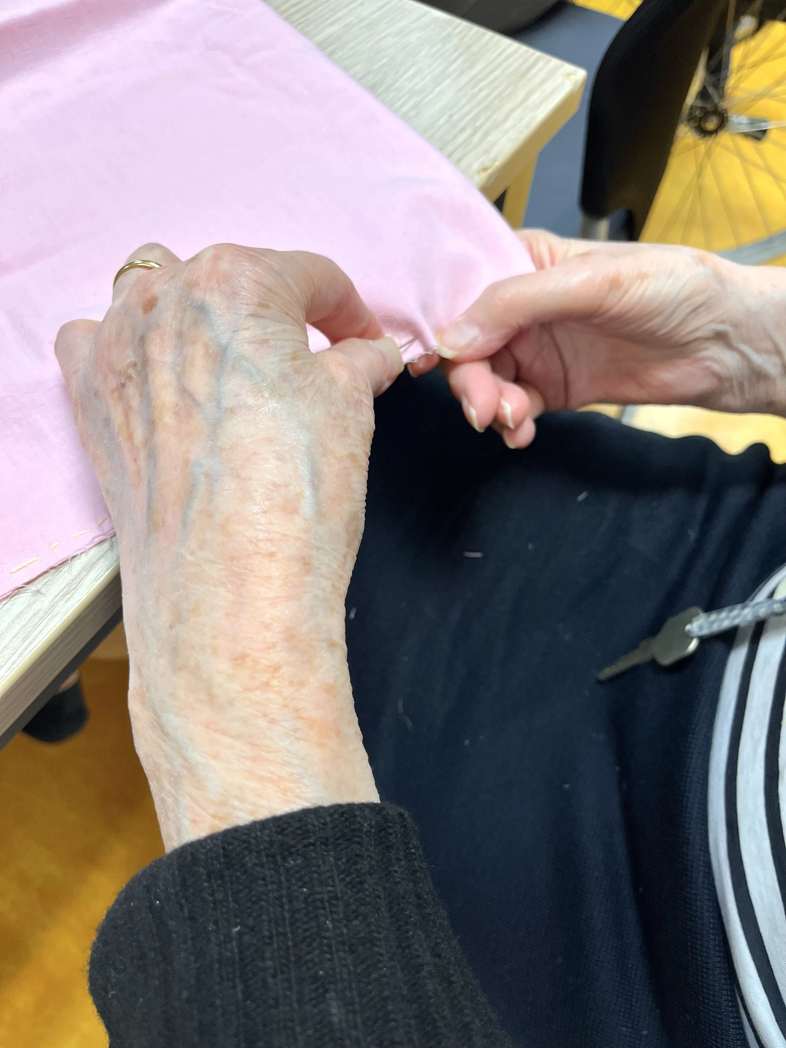 Mains de personnes âgées en train de confectionner un ouvrage en tissu rose sur une table, avec un fauteuil en arrière-plan.