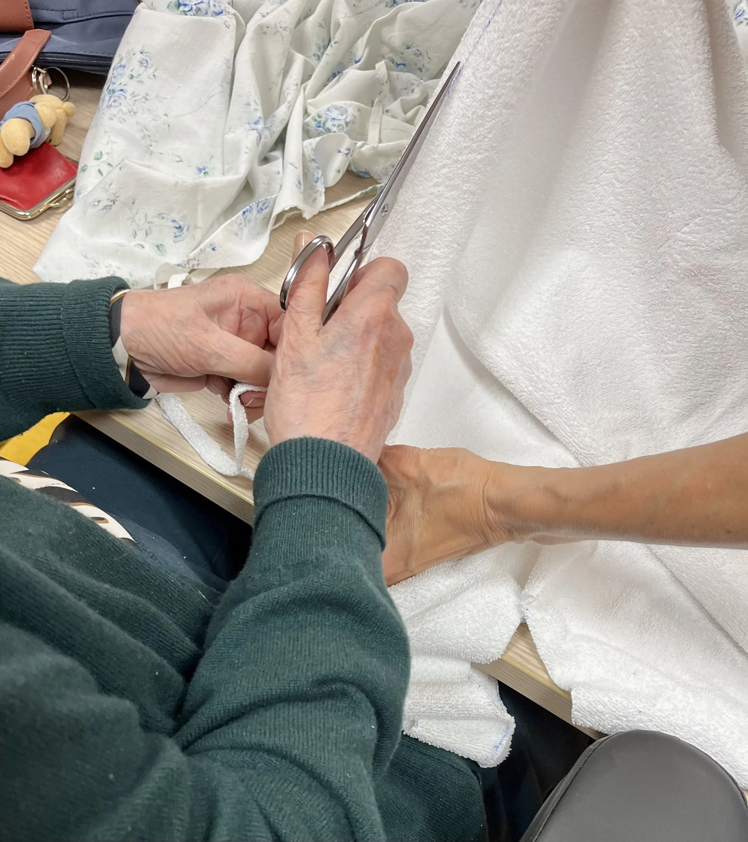 Une personne tenant une paire de ciseaux coupe un tissu blanc, tandis qu'une autre personne maintient le tissu en place. Sur la table, il y a des accessoires et des vêtements, notamment une serviette en tissu blanc, une petite figurine en peluche et un sac chemise en cuir marron.