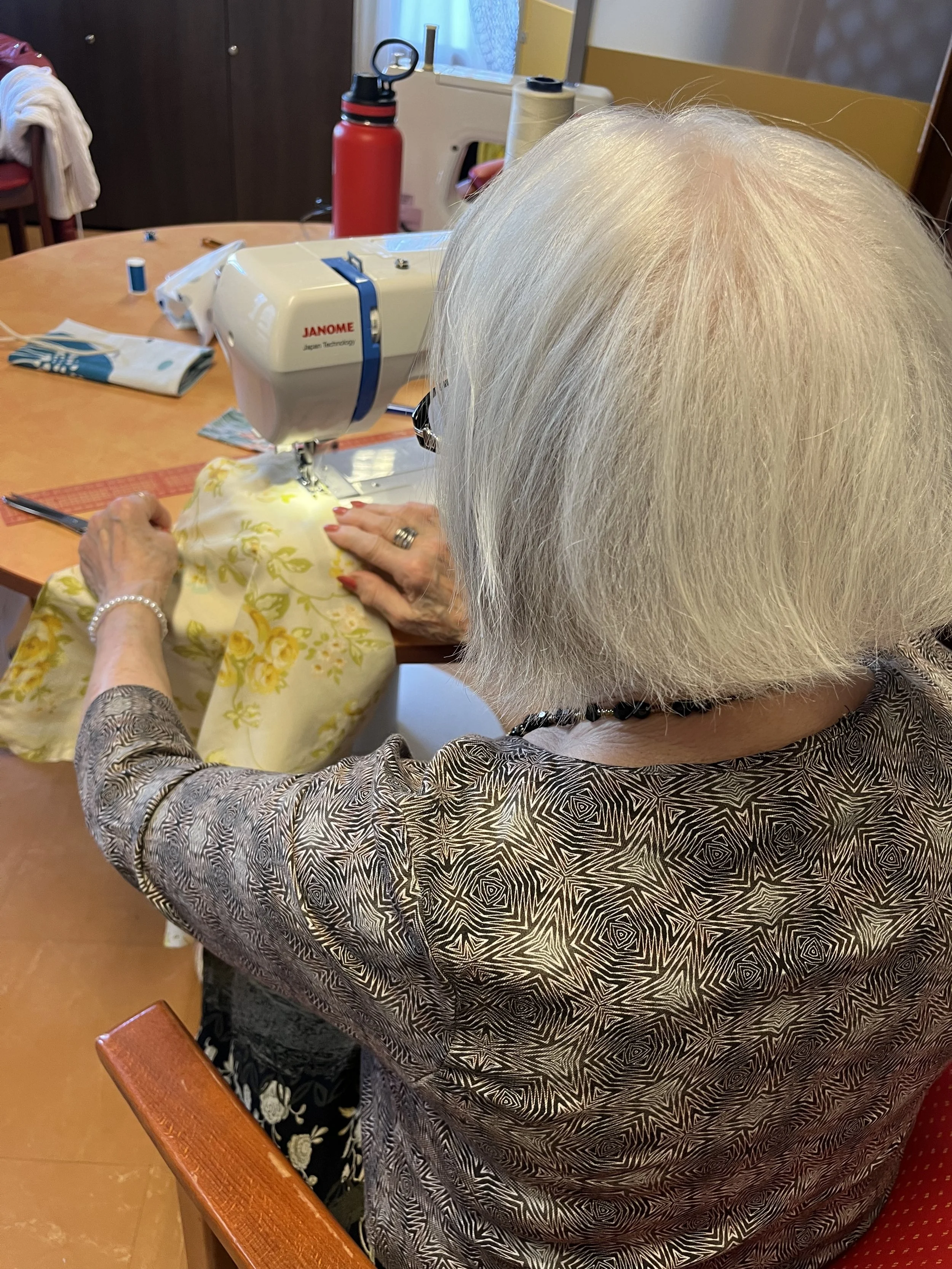 Une personne âgée avec des cheveux blancs longs cousant à une machine à coudre sur une table en bois. Autres objets de couture visibles, comme du fil et un bouton.
