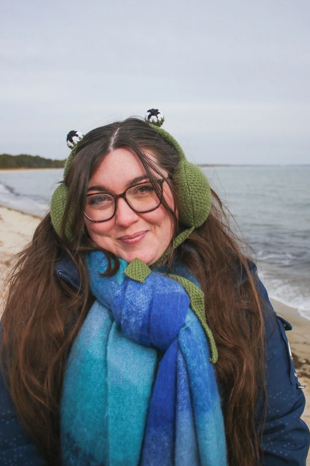Woman with glasses and long brown hair smiling on a beach, wearing green ear warmers and a blue scarf, with an overcast sky and ocean in the background.