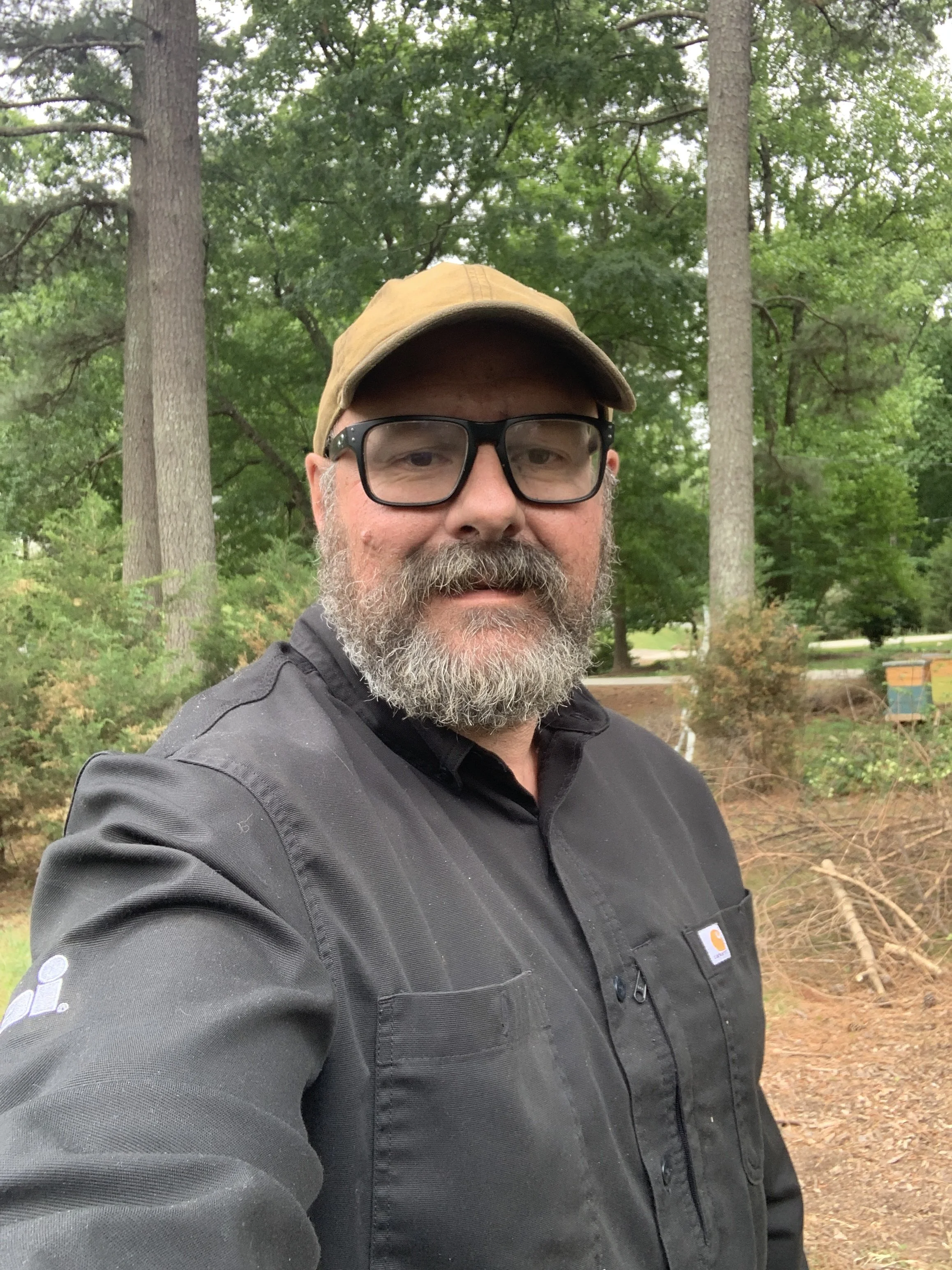 A man with glasses and a beard wearing a tan cap and a black Carhartt jacket taking a selfie outdoors in a wooded area with green trees and bushes in the background.
