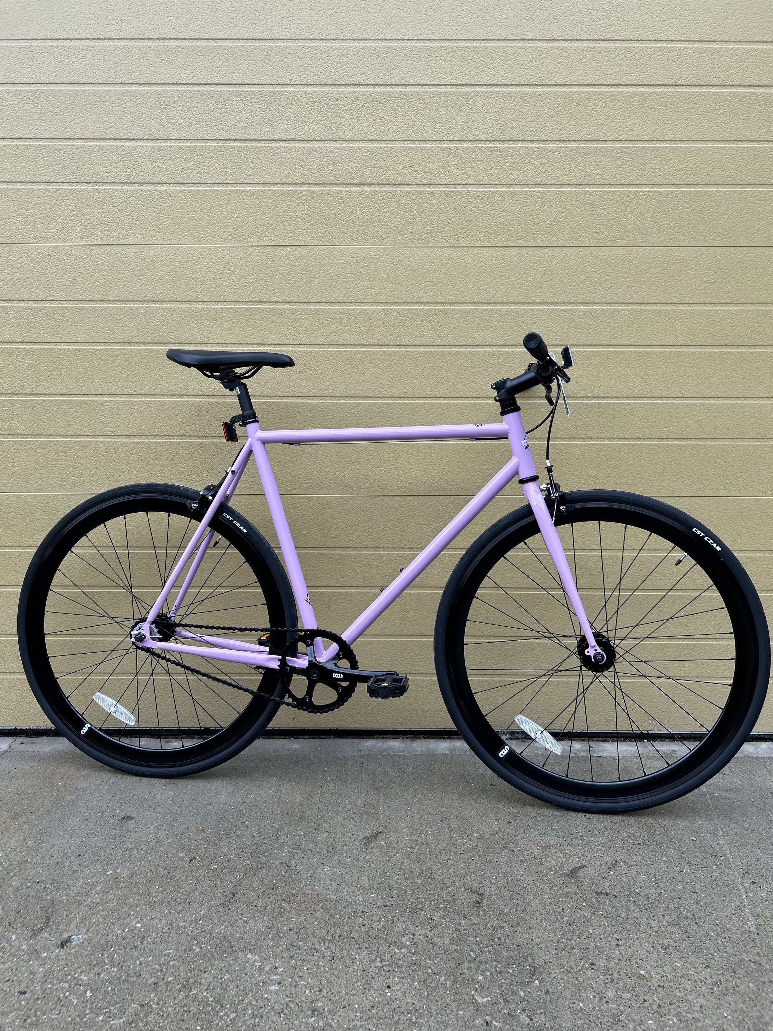 A light purple bicycle with black wheels, seat, and tires parked against a beige siding wall, on a concrete ground.