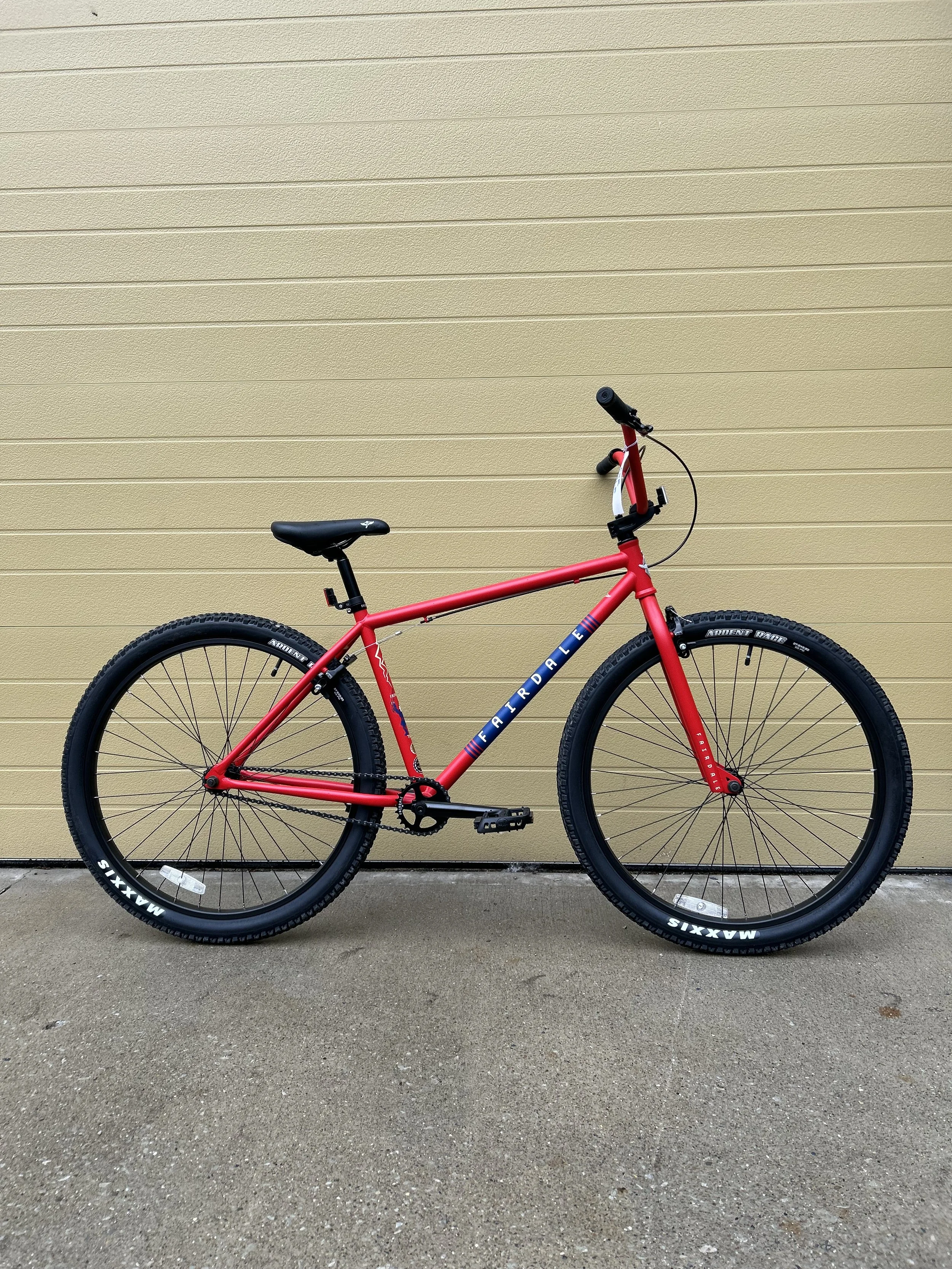 A red mountain bike with black tires and a black seat is parked against a beige wall on a concrete ground.
