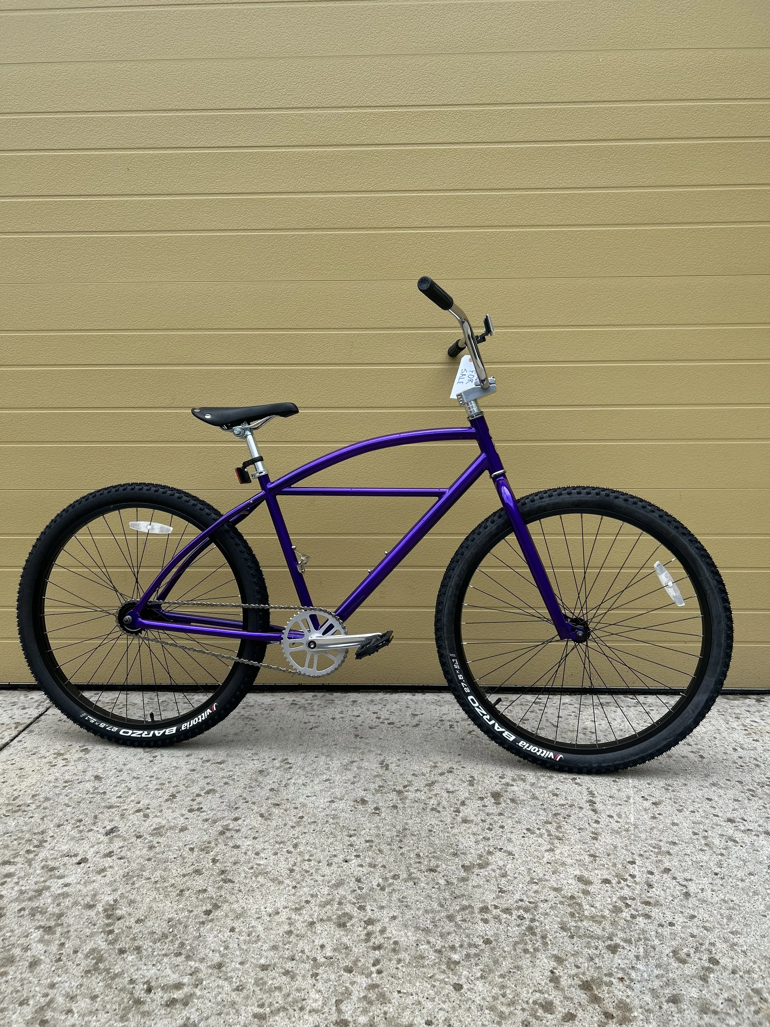 A purple mountain bike with black tires and silver pedals parked on a concrete sidewalk in front of a yellow wall.