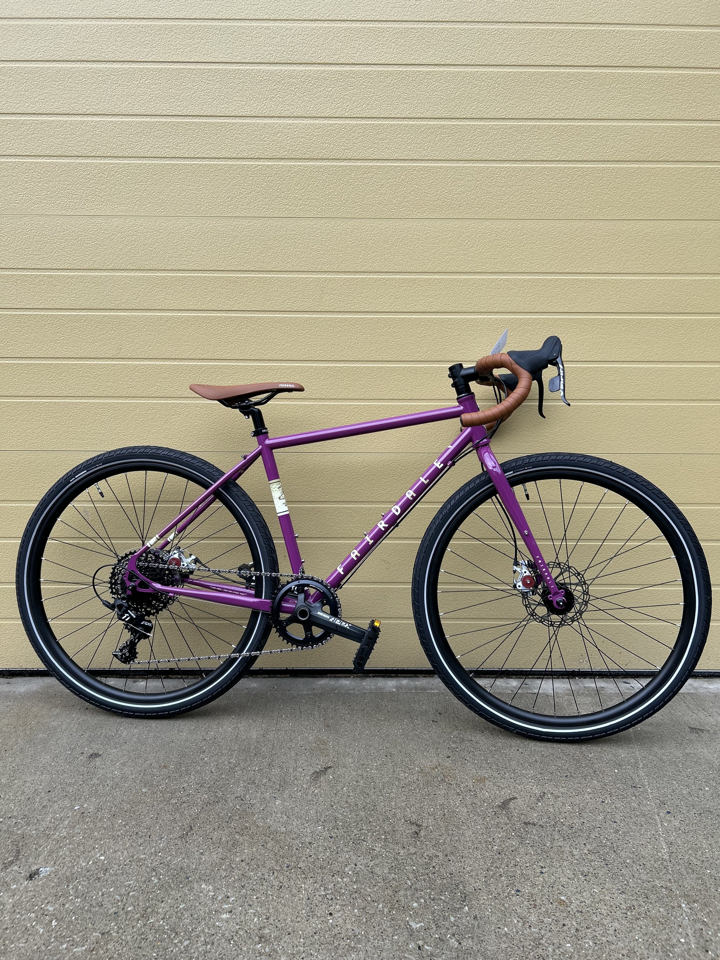 A pink and purple bicycle with a brown saddle and handlebar grips, parked against a beige paneled wall on a concrete ground.