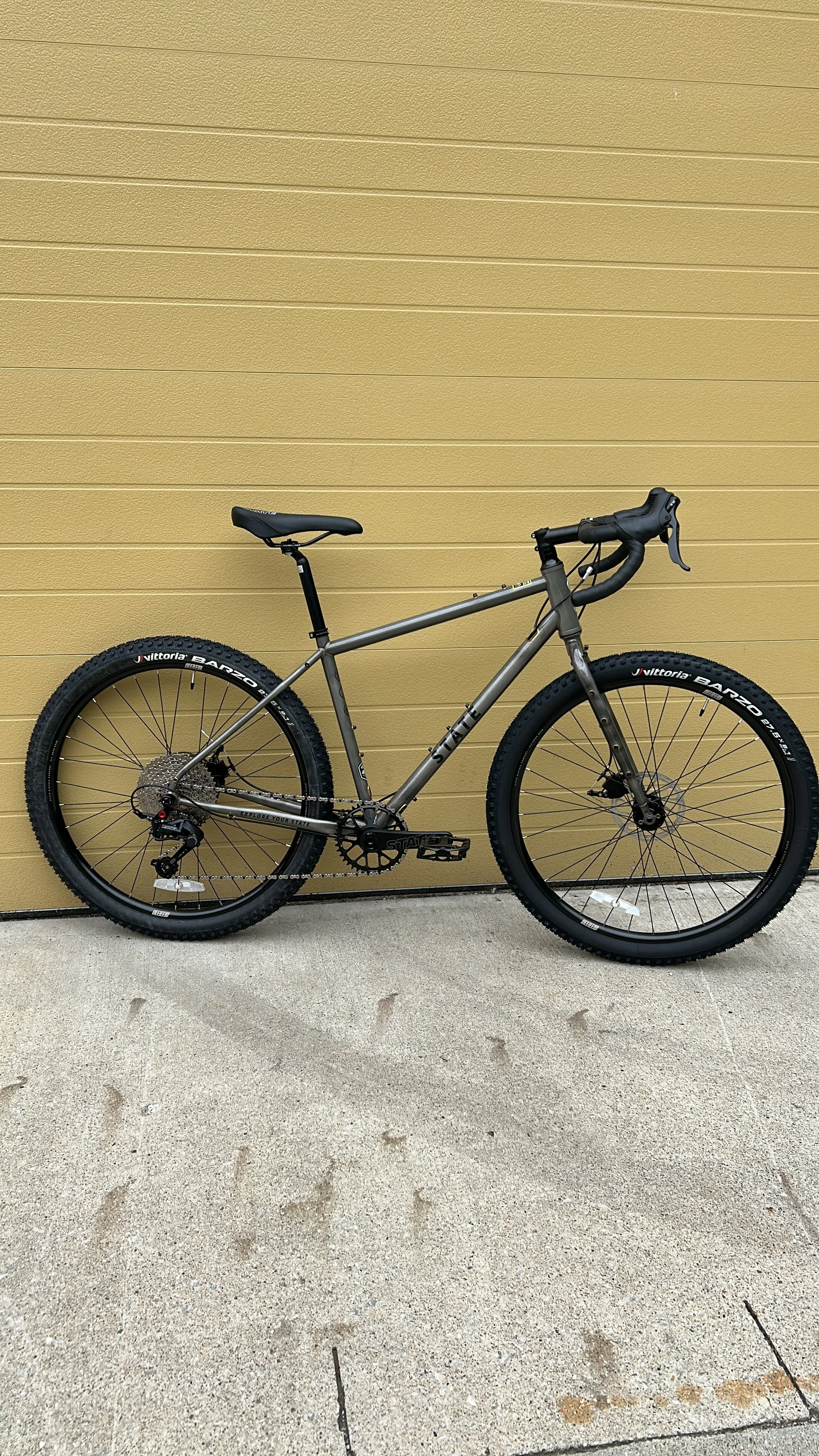 A silver mountain bike with black tires and a black saddle, parked against a beige wall on a concrete surface.
