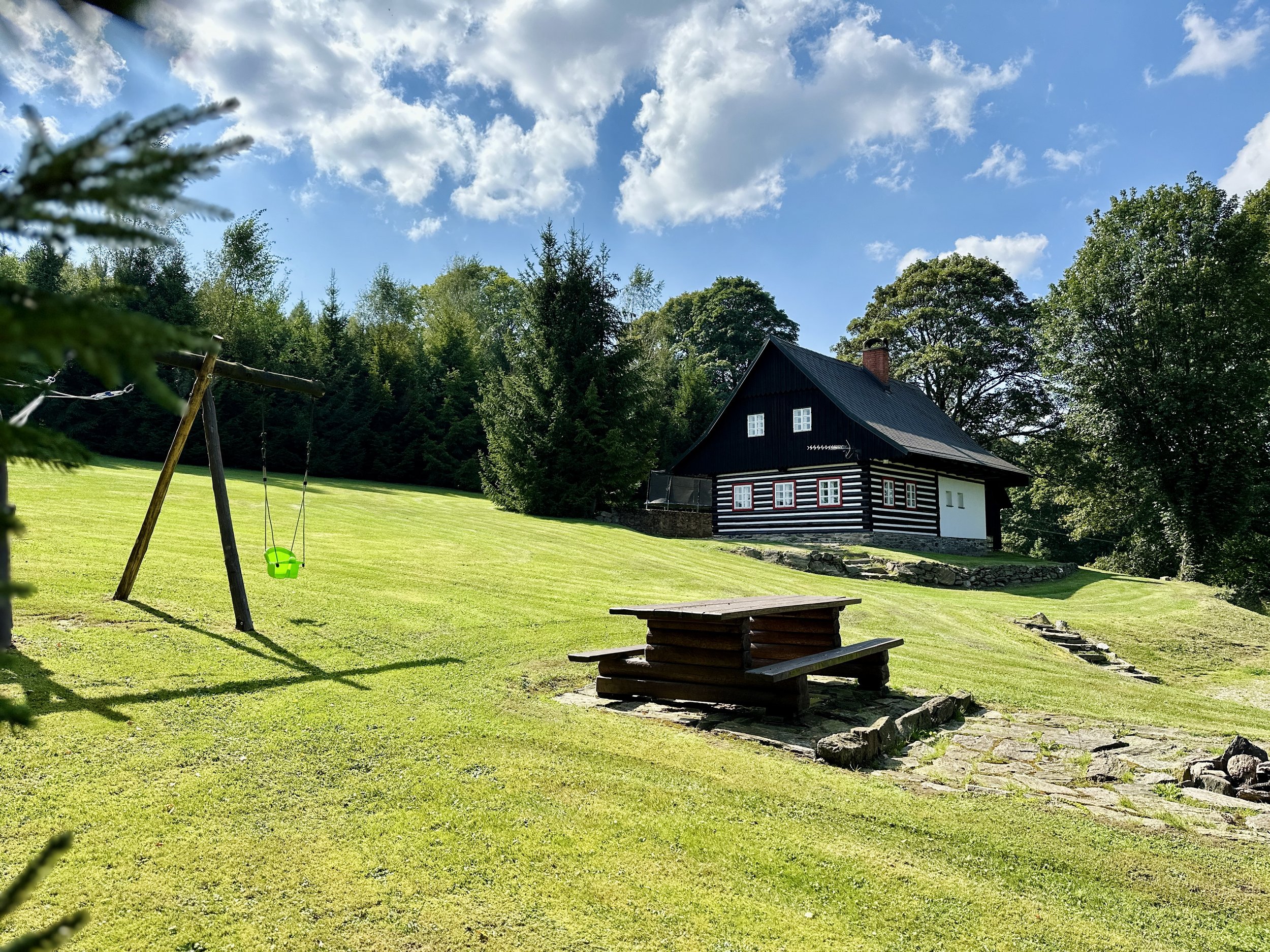 A backyard with a green lawn, a wooden swing set with a green seat, a black-and-white wooden house with red and white windows, surrounded by trees under a partly cloudy blue sky.