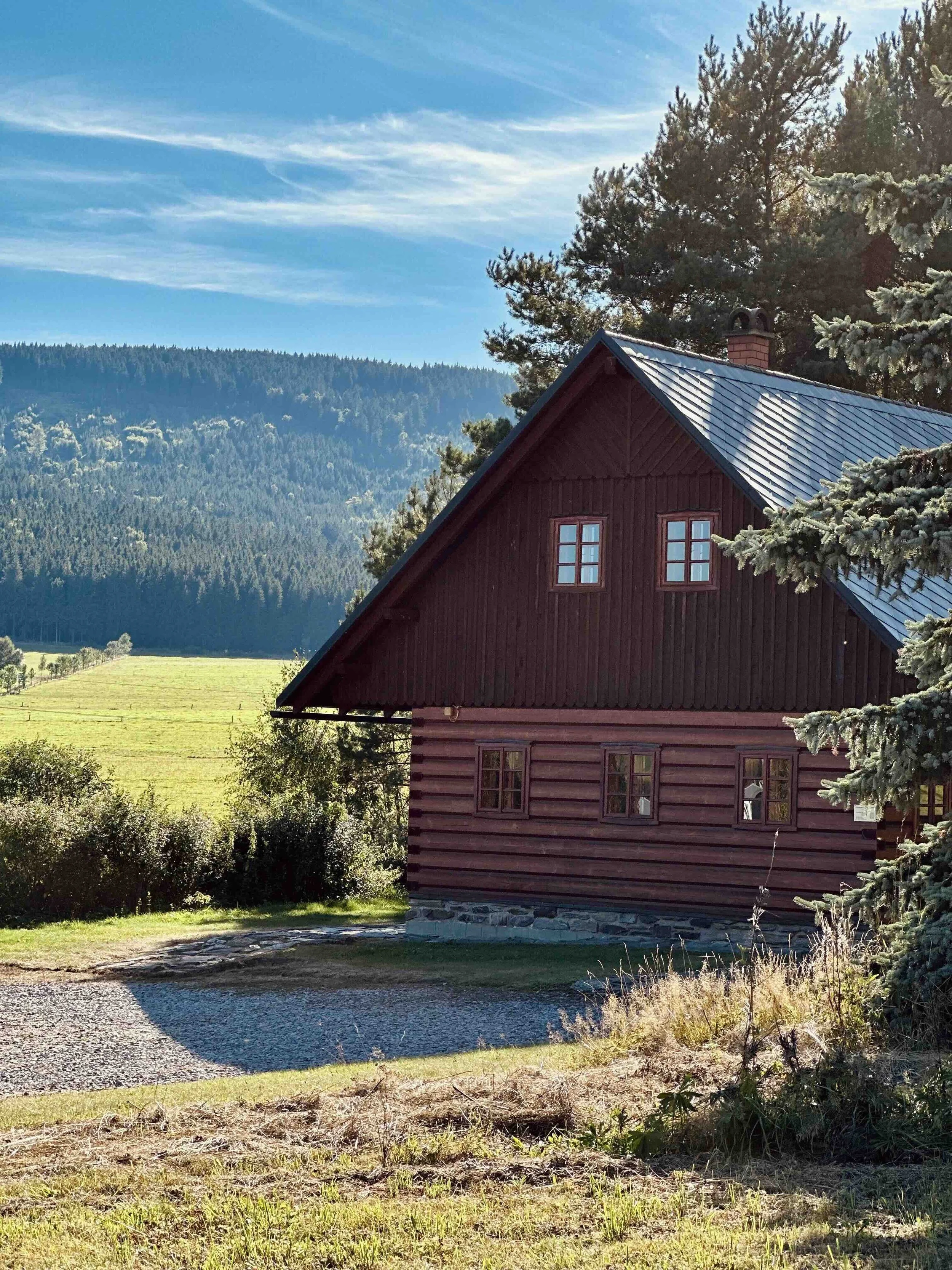 A red wooden house with a metallic roof, set against a scenic backdrop of forested hills and a blue sky with scattered clouds.