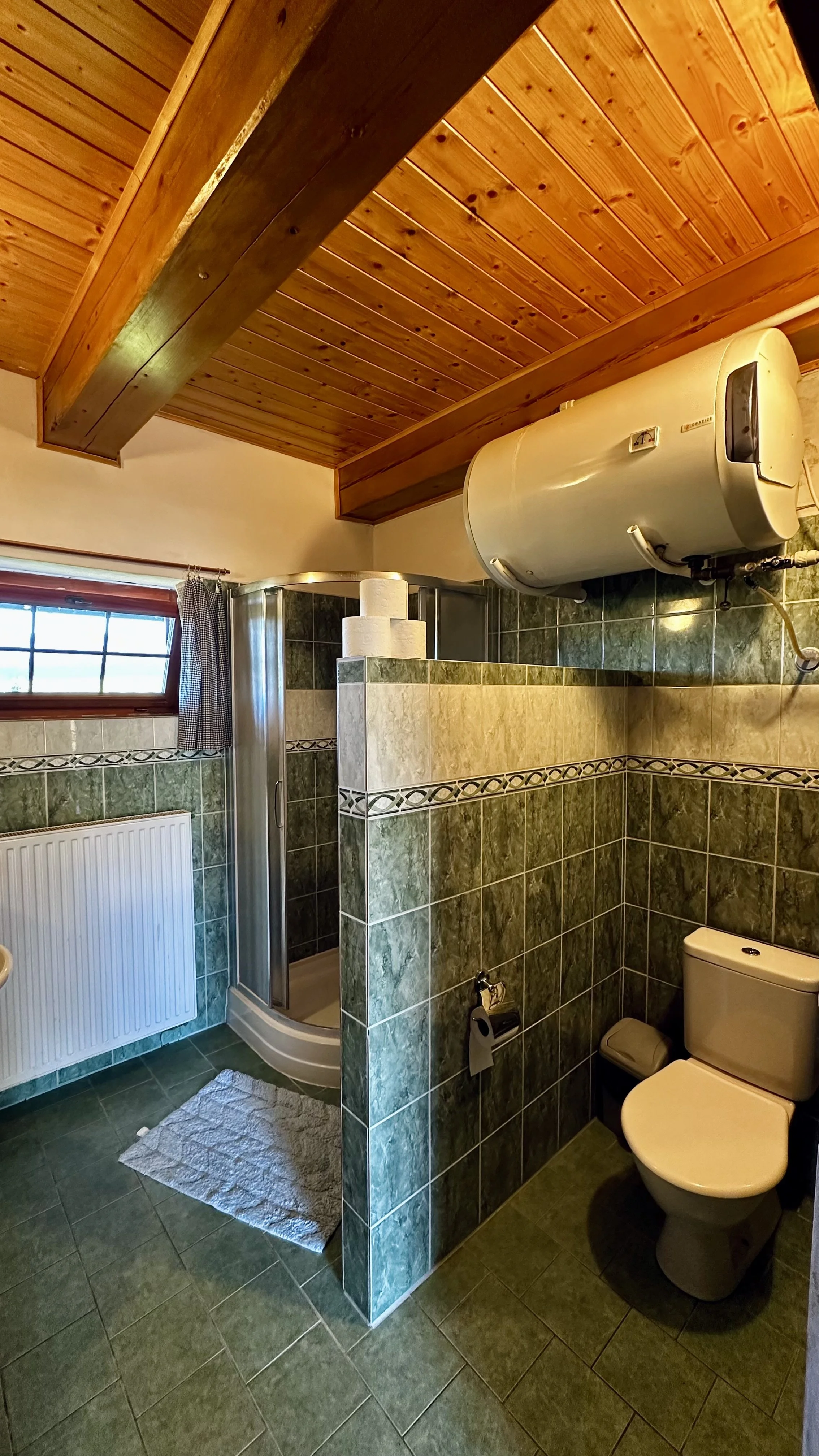 A small bathroom with green tiled floors and walls, a toilet, a radiator, a window with a small curtain, a shower enclosure, and a water heater mounted on the wall. The ceiling is wooden paneling with exposed beams, and there is a white bath mat on t
