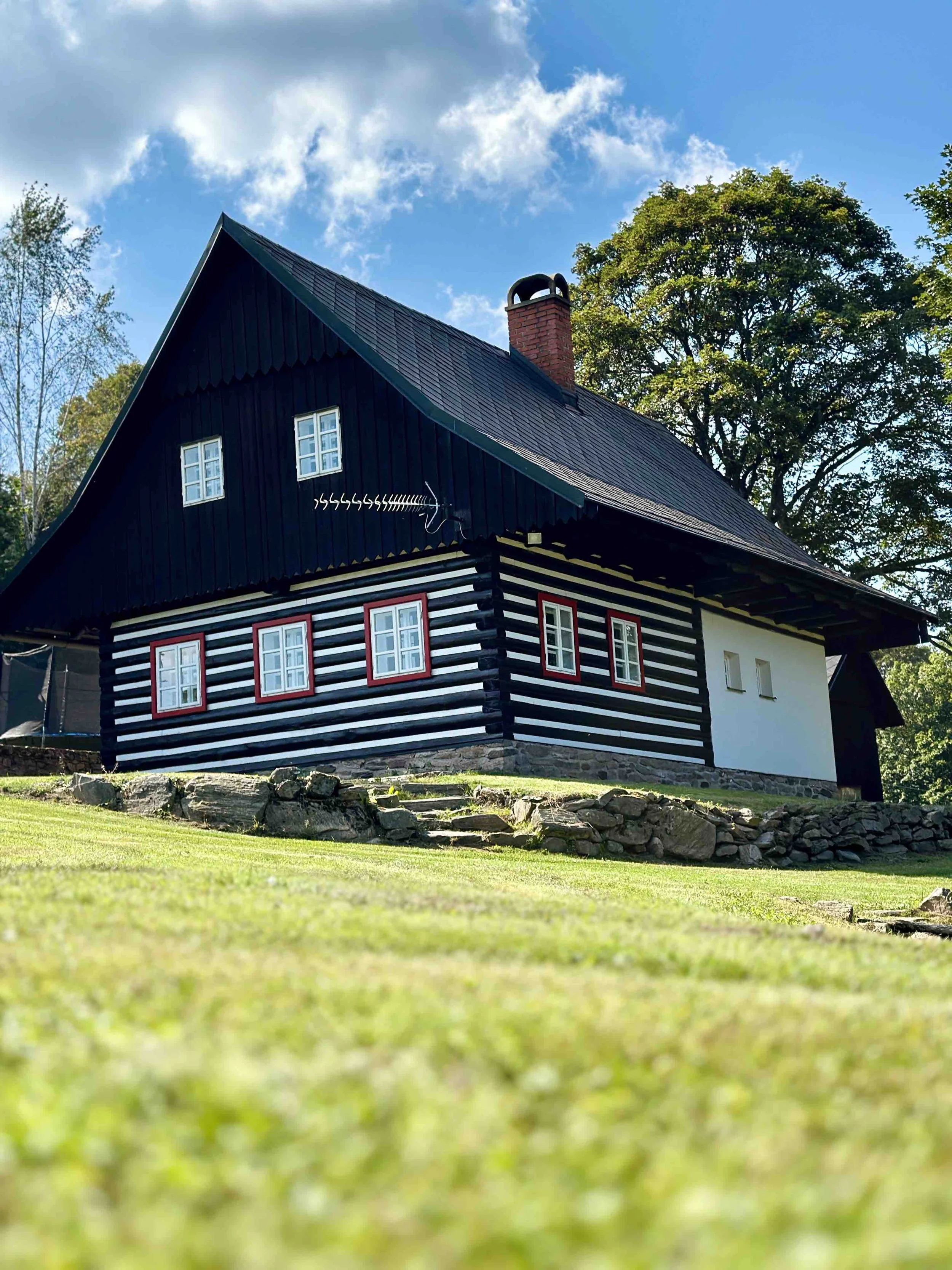 A traditional house with dark wood siding accented by red window frames, situated on a stone foundation. The house has a steeply pitched roof with black shingles and a brick chimney. Surrounding trees and a clear blue sky with some clouds are visible in the background.