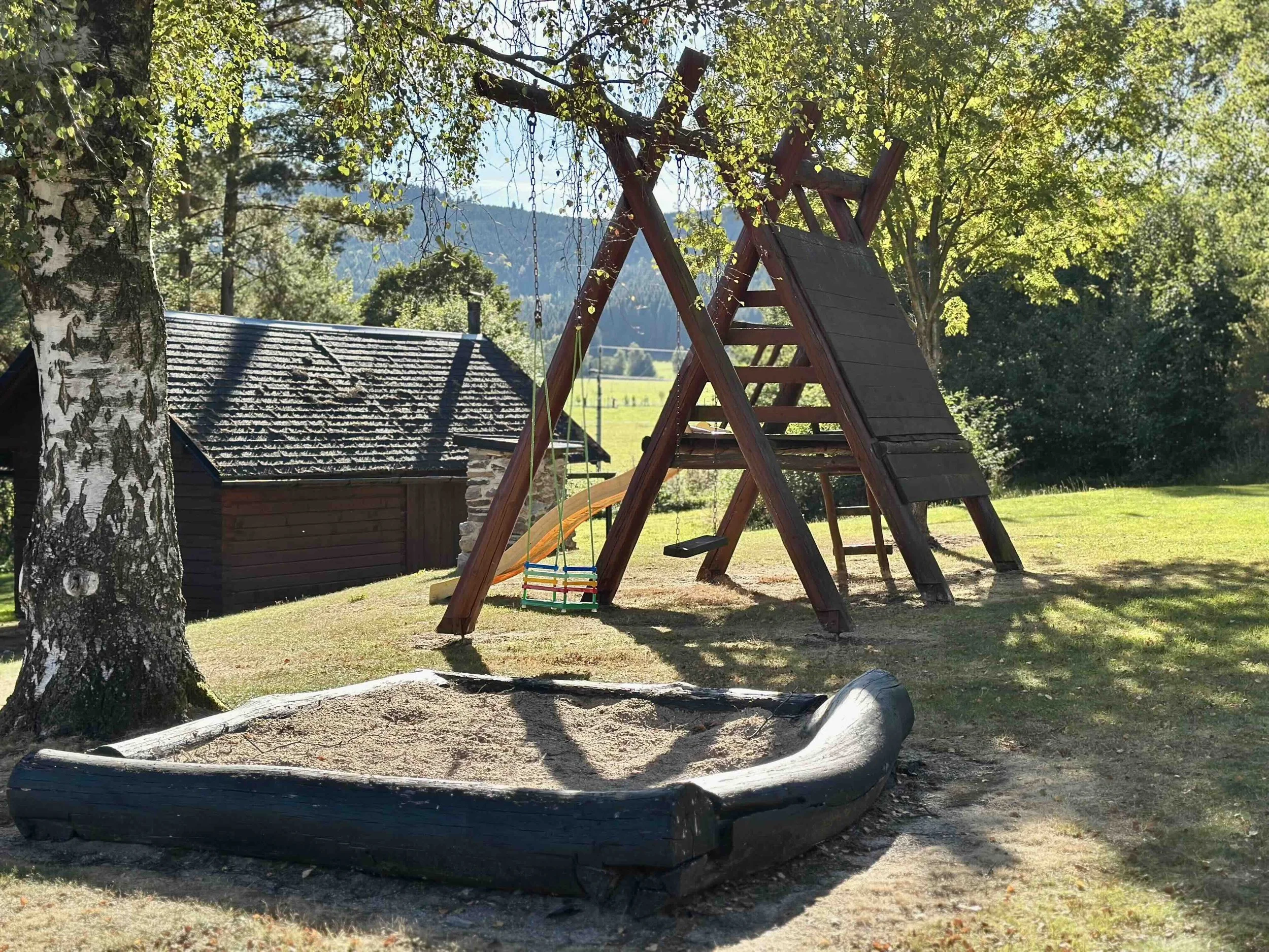 Wooden playground swing set with a slide in a grassy outdoor area surrounded by trees and a building nearby.